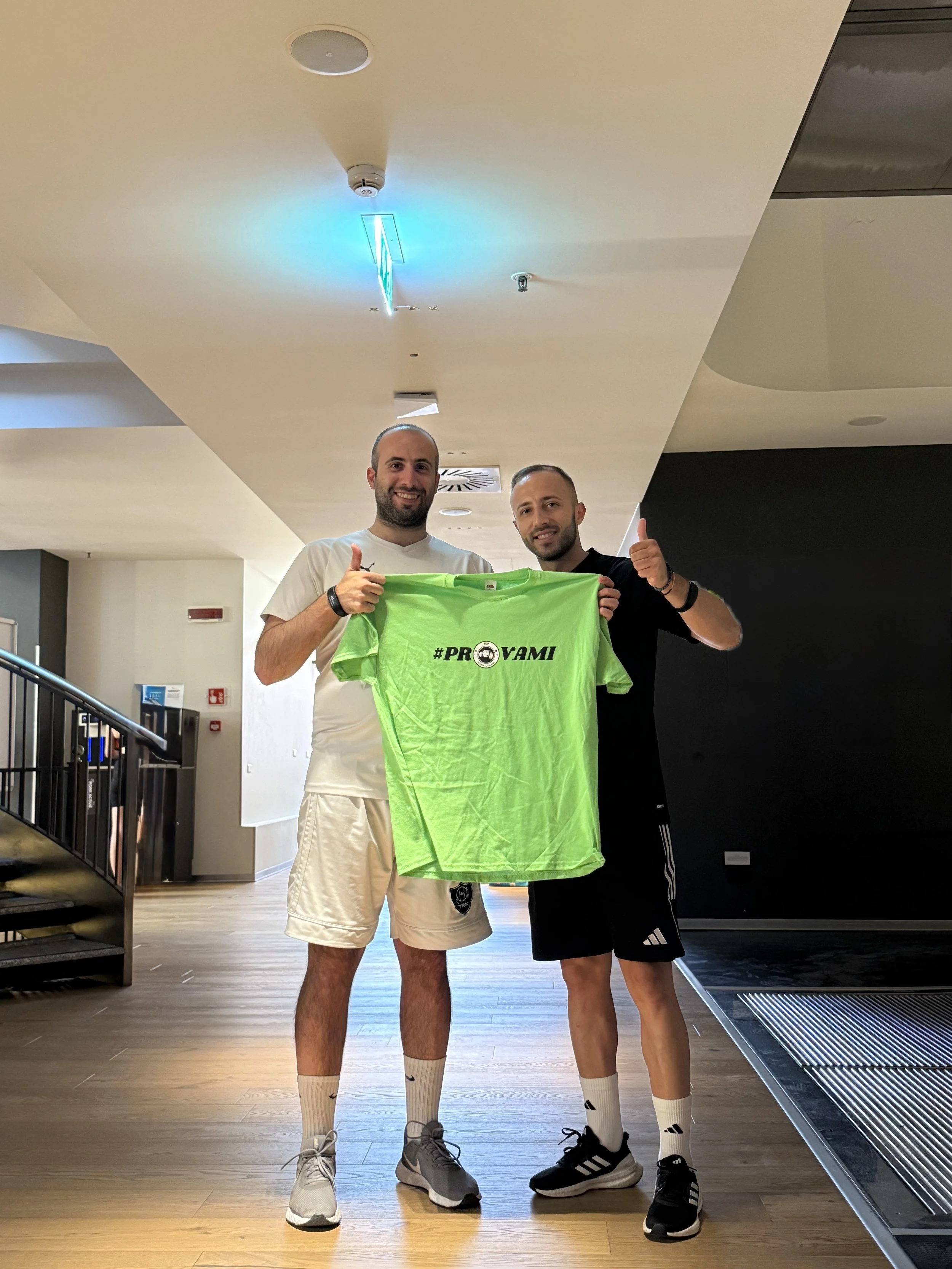 Due uomini felici in abbigliamento sportivo sorridono, uno dei due mostra una maglietta verde con la scritta '#PROVAMÌ' in un ambiente interno.