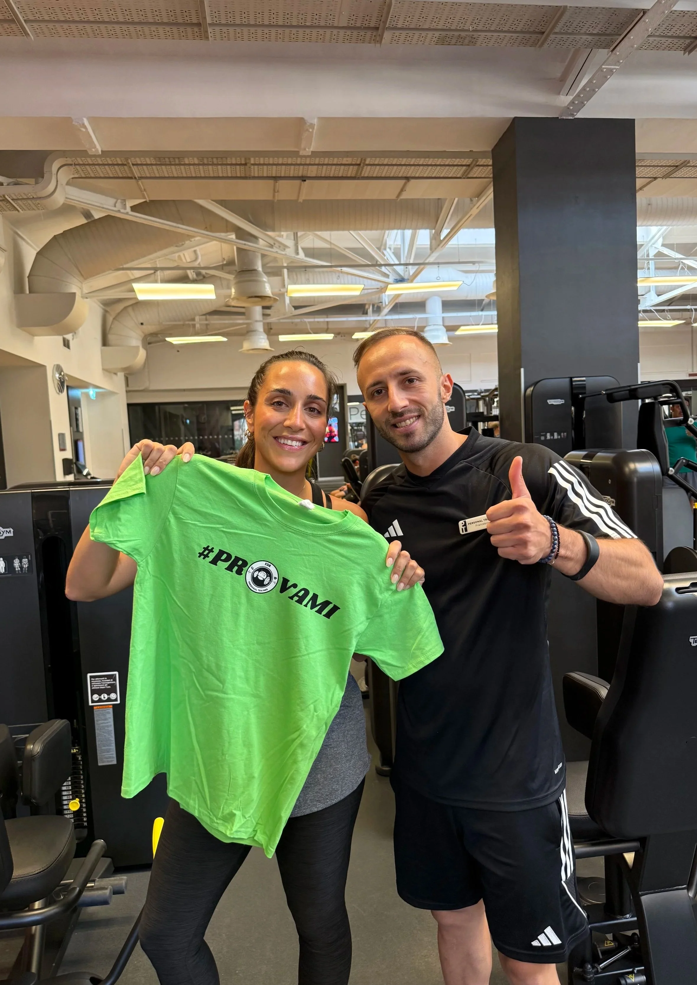 Due persone sorridenti in palestra, una donna e un uomo, tengono una maglietta verde con scritto #PROVAMI, mentre fanno il gesto del pollice in su.