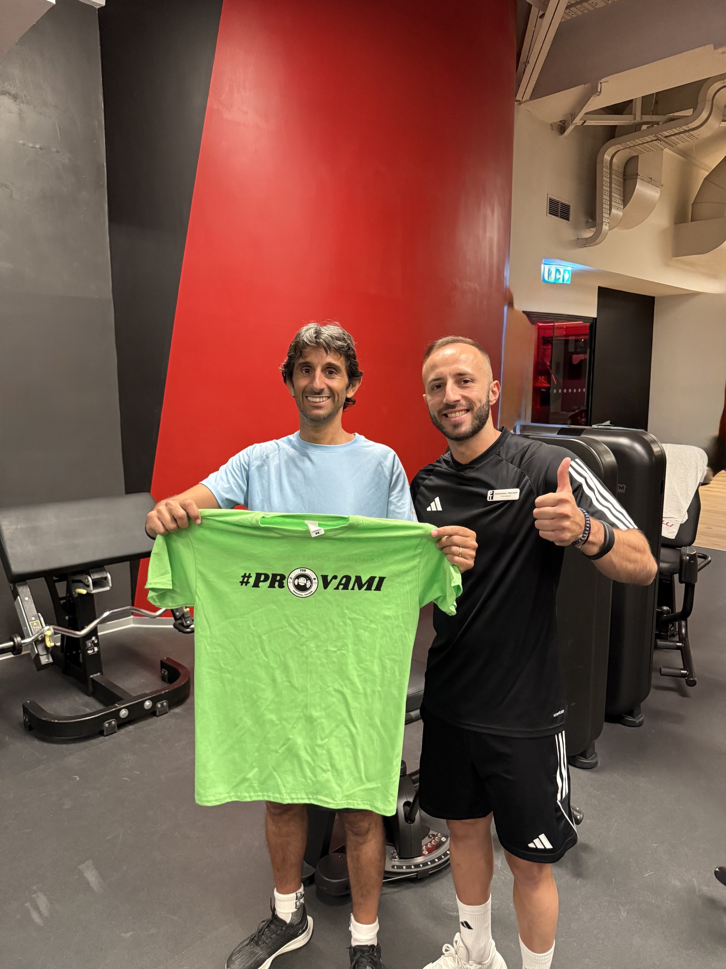 Due uomini sorridenti in palestra, uno tiene una maglietta verde con la scritta #PROVAMI e un'icona di macchina fotografica, l'altro fa il pollice alzato.