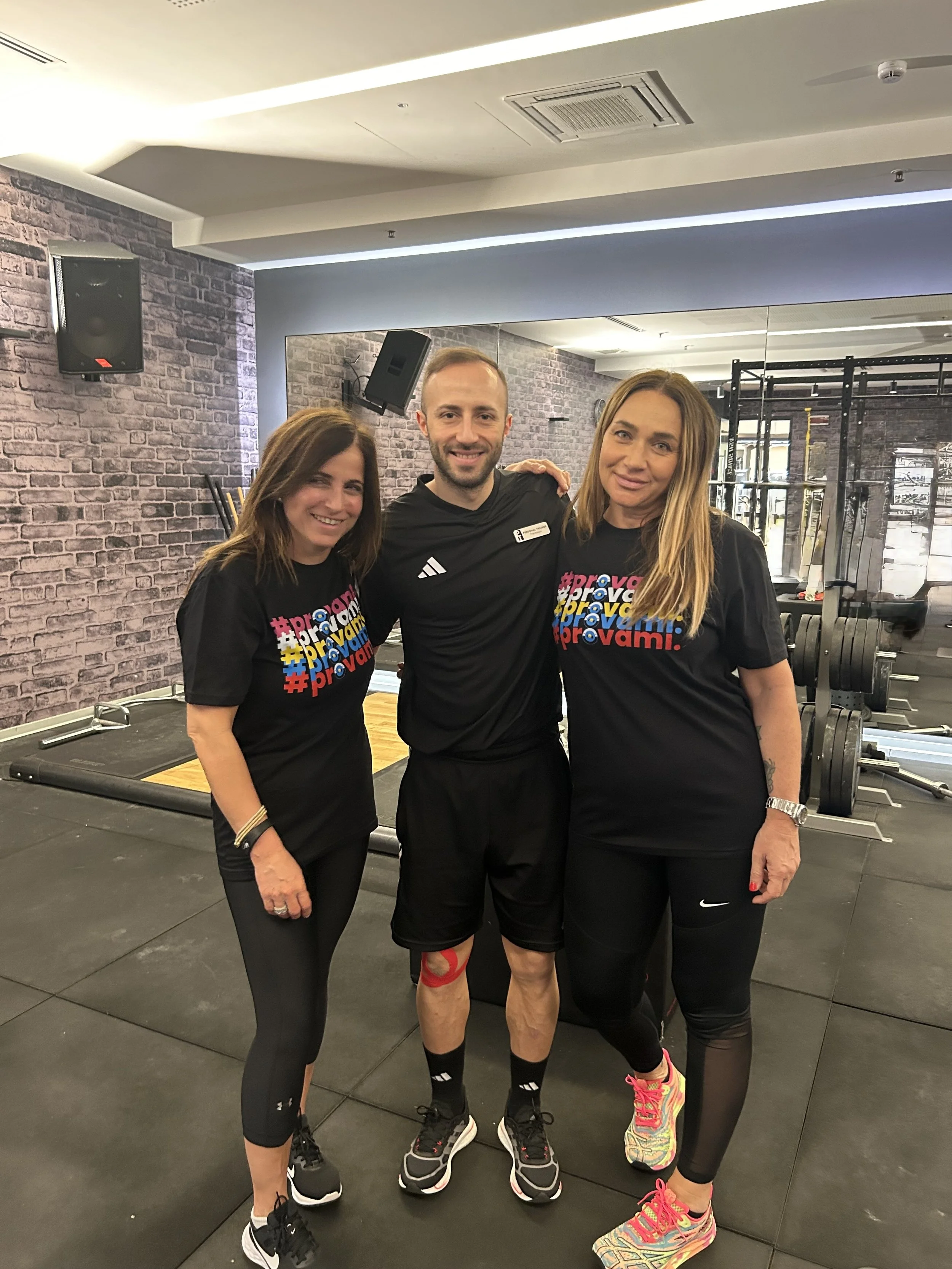 Tre persone sorridenti in palestra, due donne e un uomo, indossano abbigliamento sportivo e si abbracciano, con attrezzature per allenamento sullo sfondo.