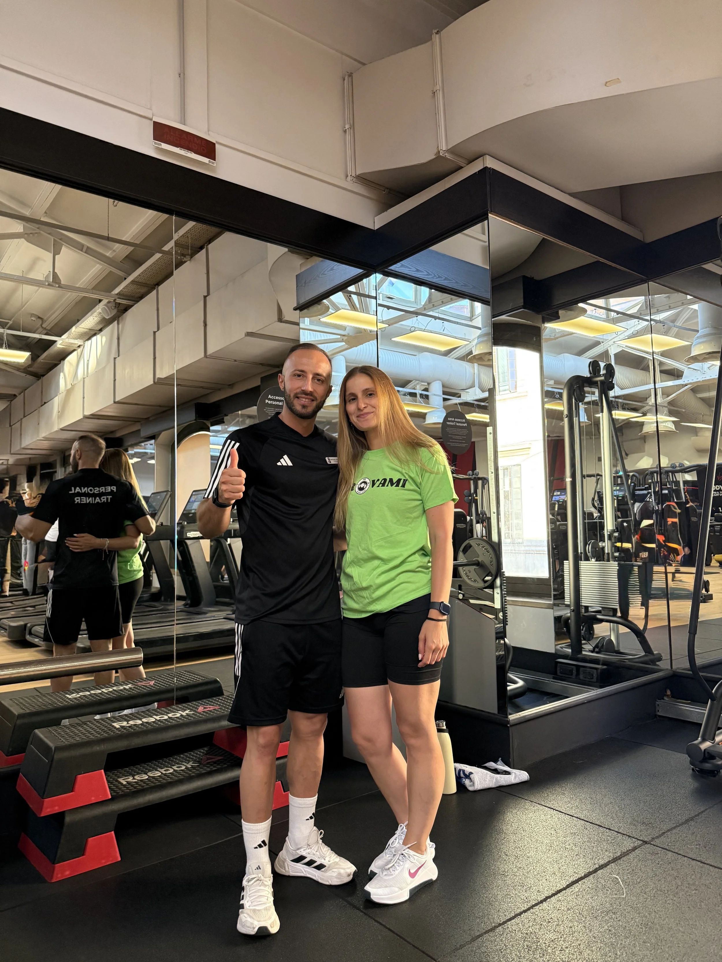 Due persone felici in palestra, un uomo con maglietta nera e donna con maglietta verde, posa sorridendo.