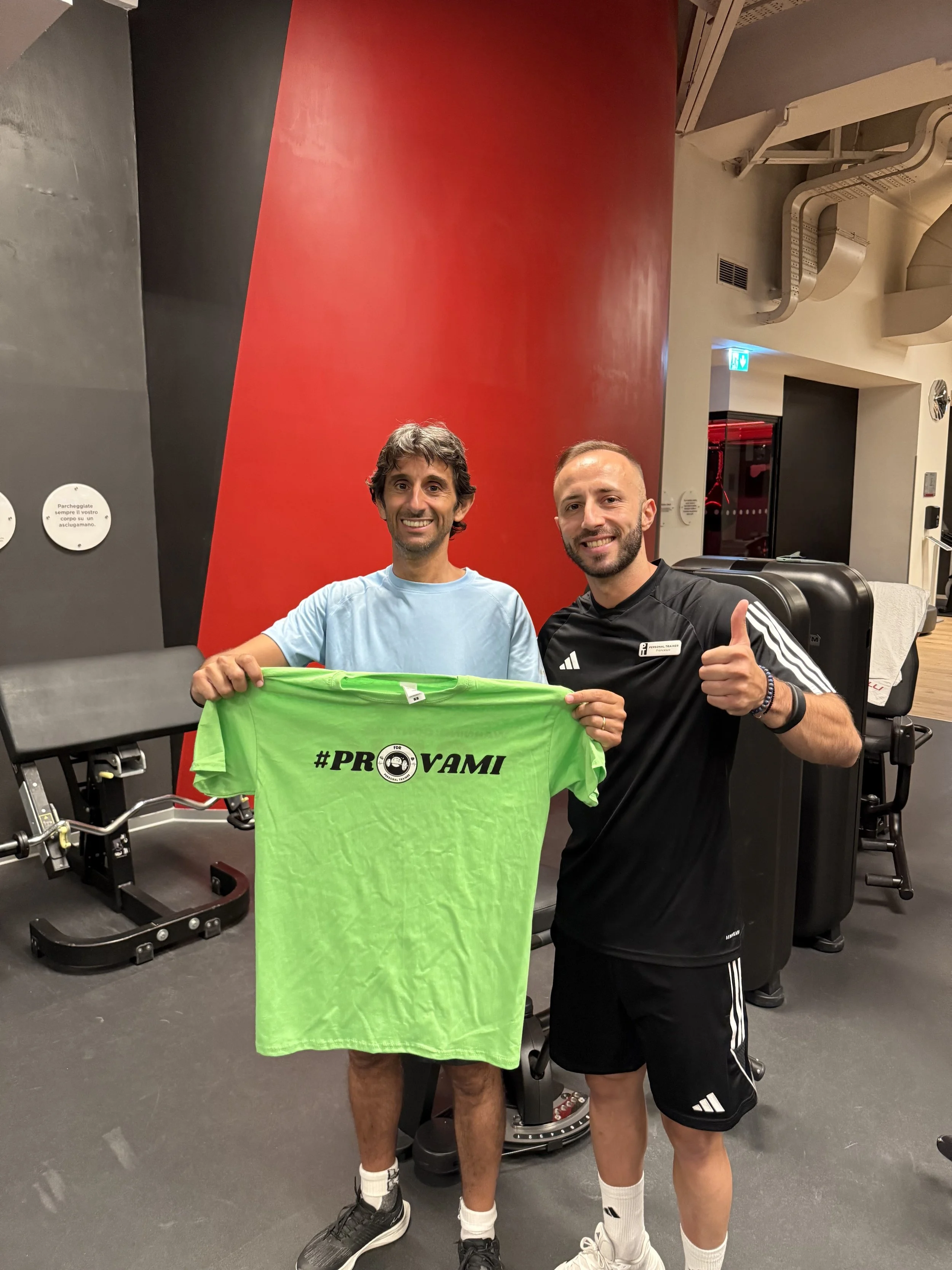 Due uomini sorridenti in palestra, uno tiene una maglietta verde con la scritta #PROVAMI e un'icona di macchina fotografica, l'altro fa il pollice alzato.