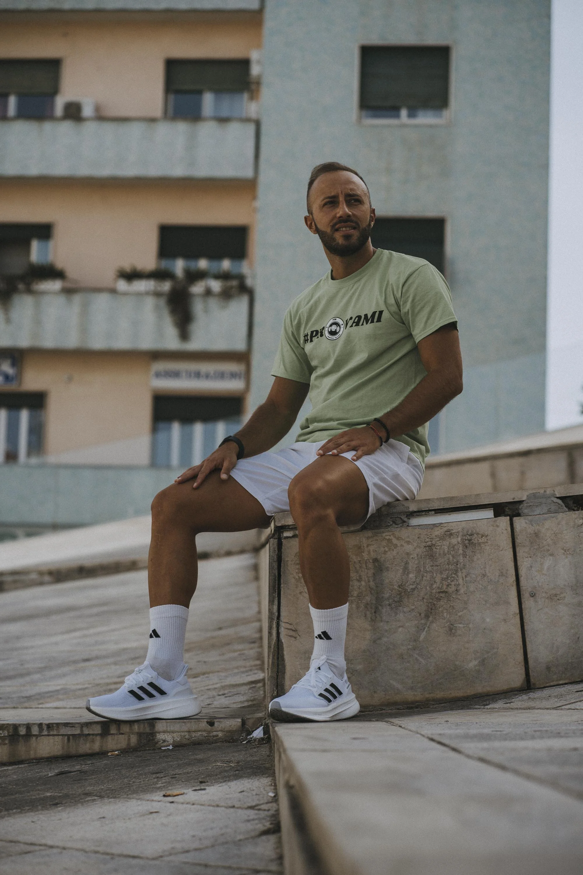 Uomo seduto su un gradino in una zona urbana, abbigliato con t-shirt verde, pantaloncini bianchi, calzini bianchi e scarpe sportive bianche con strisce nere.