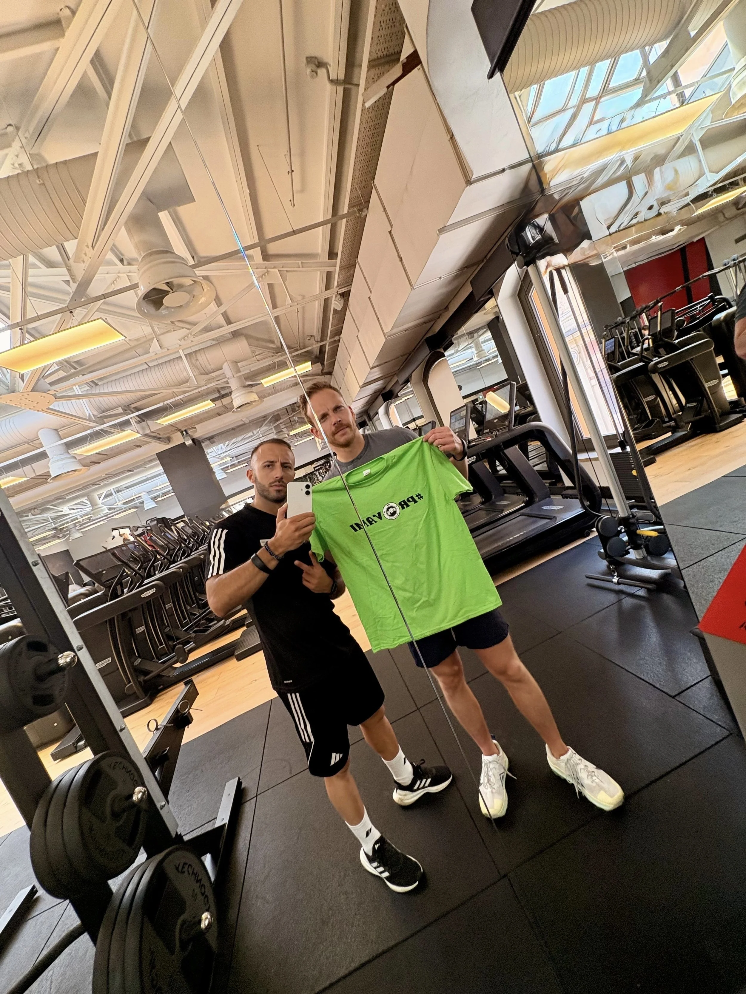 Due uomini si riflettono in uno specchio di una palestra, uno di loro tiene in mano una maglietta verde e si scatta un selfie in palestra.