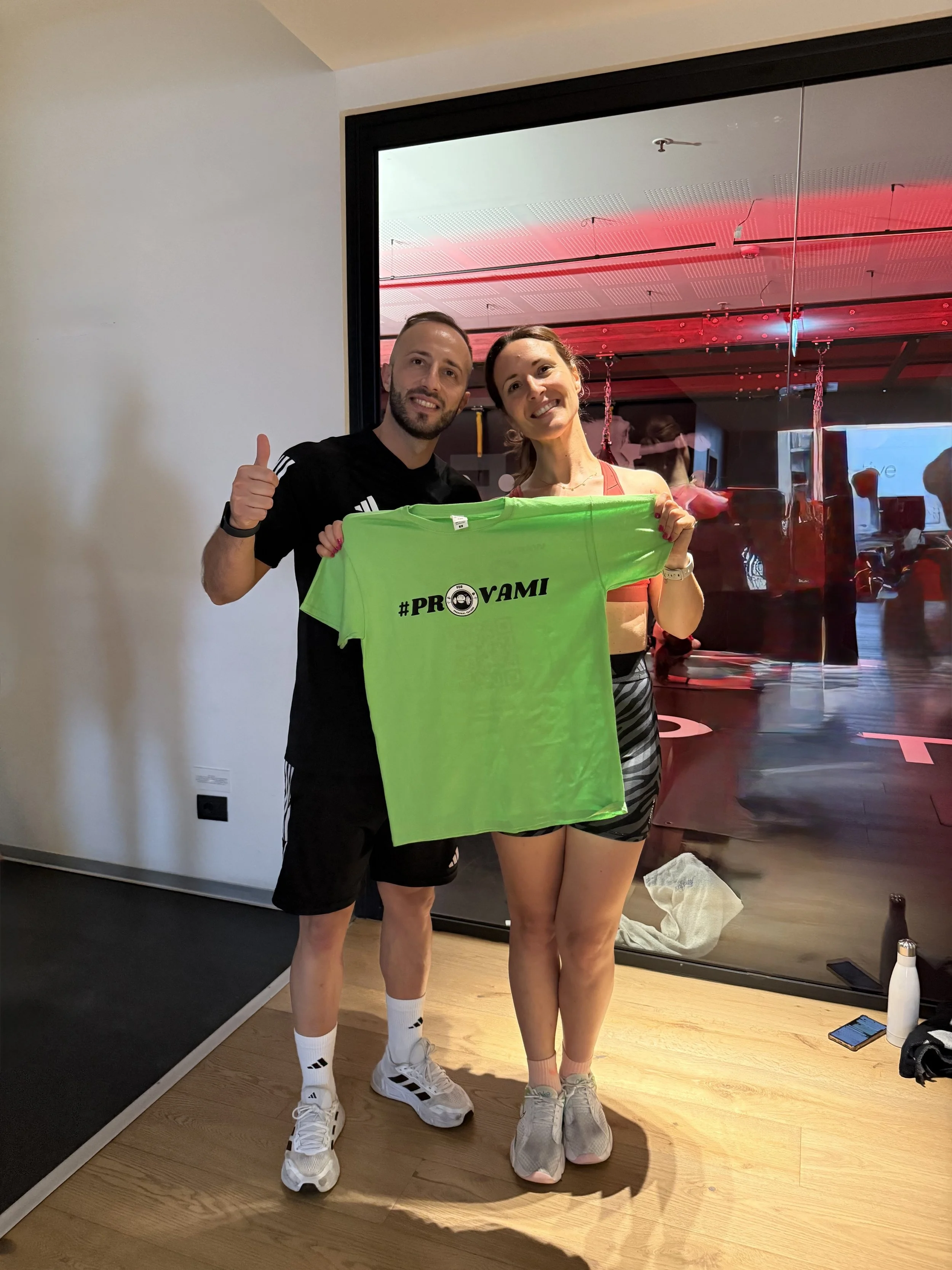 Una coppia sportiva sorridente tiene una maglietta verde con il hashtag #PROVAMI, mentre si trova in una stanza con pareti bianche e una parete di vetro che riflette luci rosse e altre persone in un'area fitness.