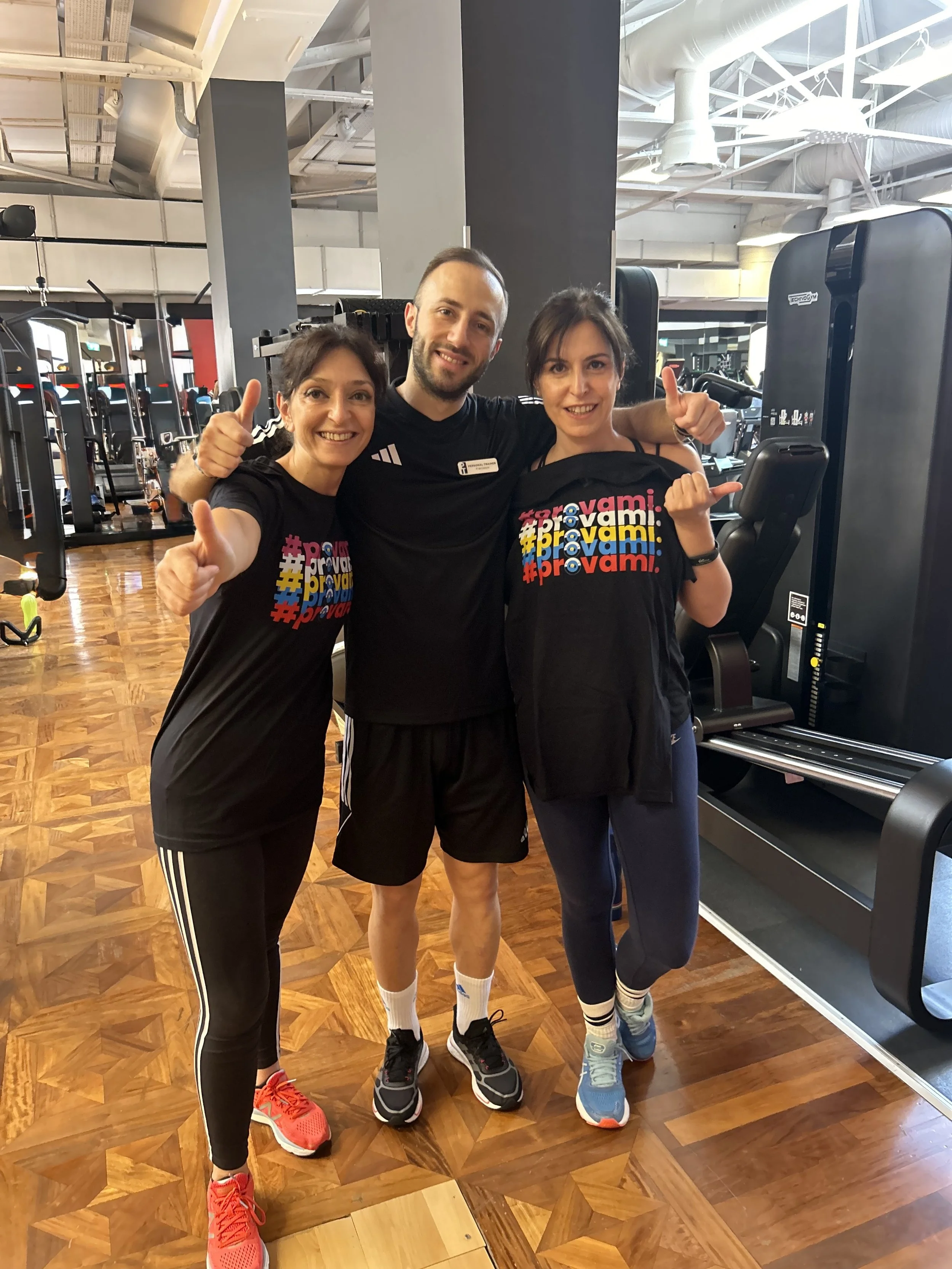Tre persone sorridenti in abbigliamento sportivo che fanno il gesto del pollice in su e il gesto della mano a forma di 'L' in un'area fitness con attrezzature da palestra in sfondo.