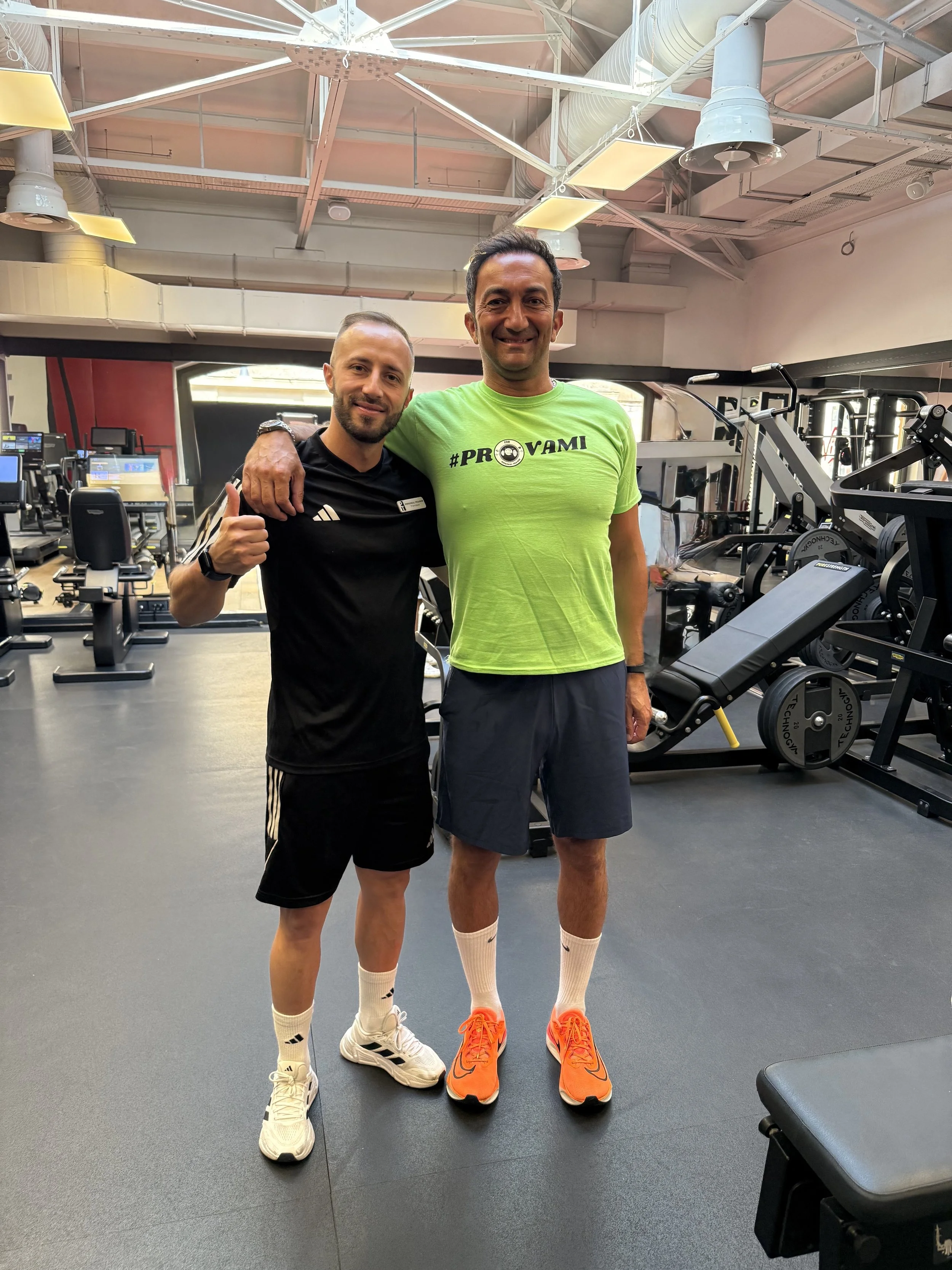 Due uomini sorridenti in palestra, uno con braccio intorno all'altro, vestiti sportivamente, in uno studio di allenamento con attrezzature da fitness visibili sullo sfondo.