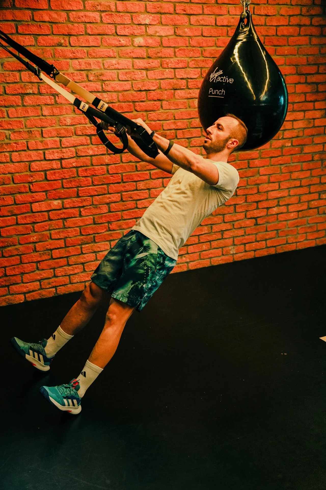 Uomo che si allena in palestra con un sacco da boxe e cinghie di resistenza, in piedi su un pavimento nero contro una parete di mattoni rossi.