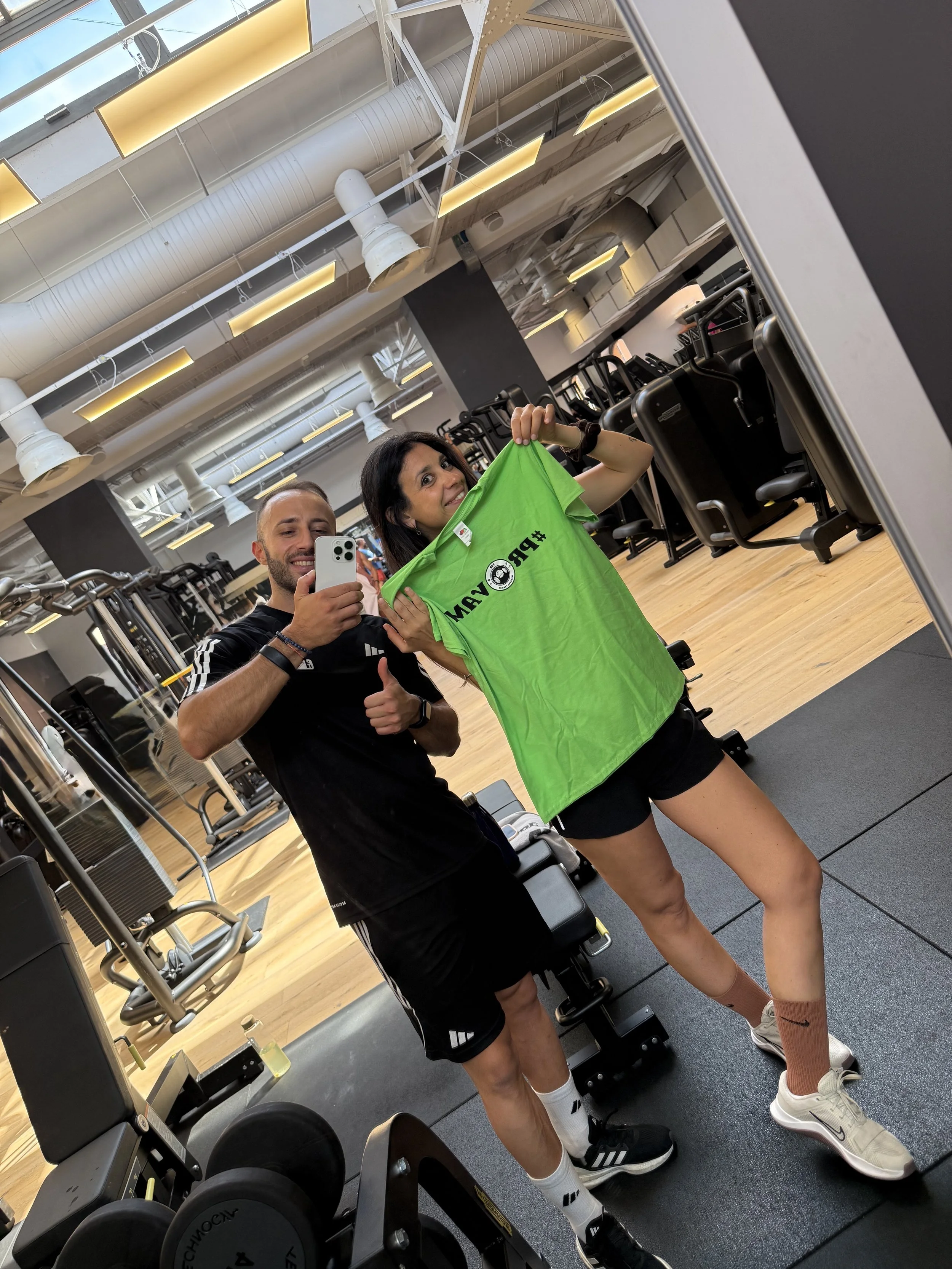 Due persone in palestra che fanno un selfie nello specchio. La donna tiene una maglietta verde con scritte e il logo di WWE, mentre l'uomo mostra il pollice verso. Entrambe sono vestite sportivamente con scarpe da ginnastica.