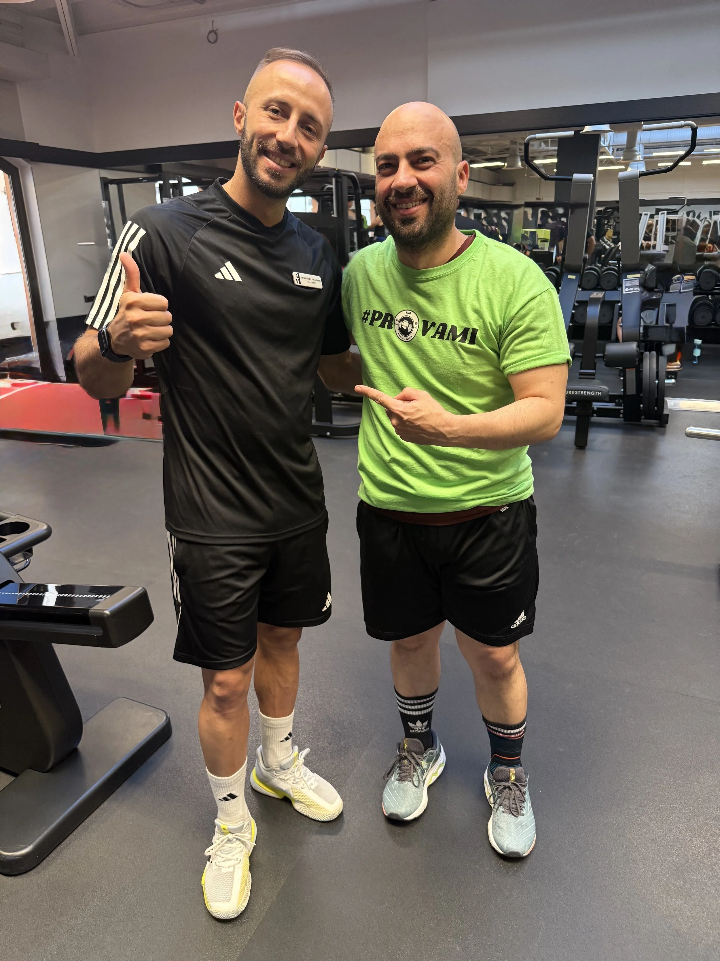 Due uomini sorridenti in abbigliamento sportivo in palestra, uno indossa una maglietta nera con loghi Adidas, l'altro una maglietta verde con la scritta #PROVAMI, uno dei due indica l'altro.