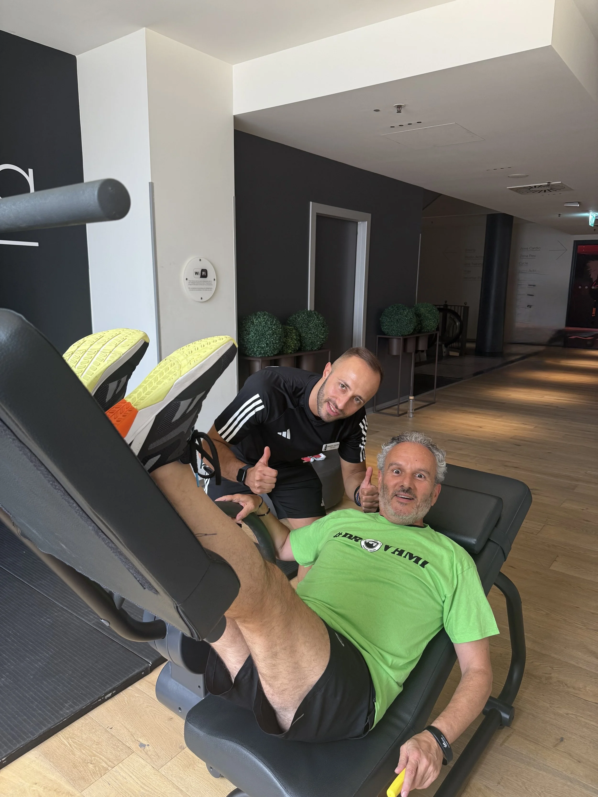 Due uomini in un centro fitness, uno sdraiato su una panca e l'altro che si trova vicino, entrambi sorridenti e che salutano con il pollice in su.