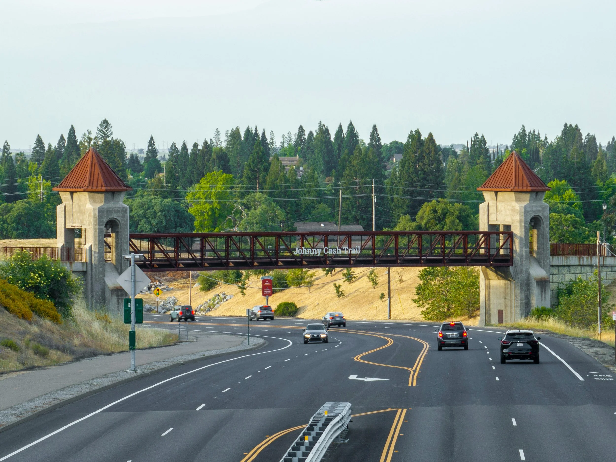 A roadway with cars passing under a pedestrian bridge labeled Johnny Cash Trail, set against a backdrop of trees and hills.