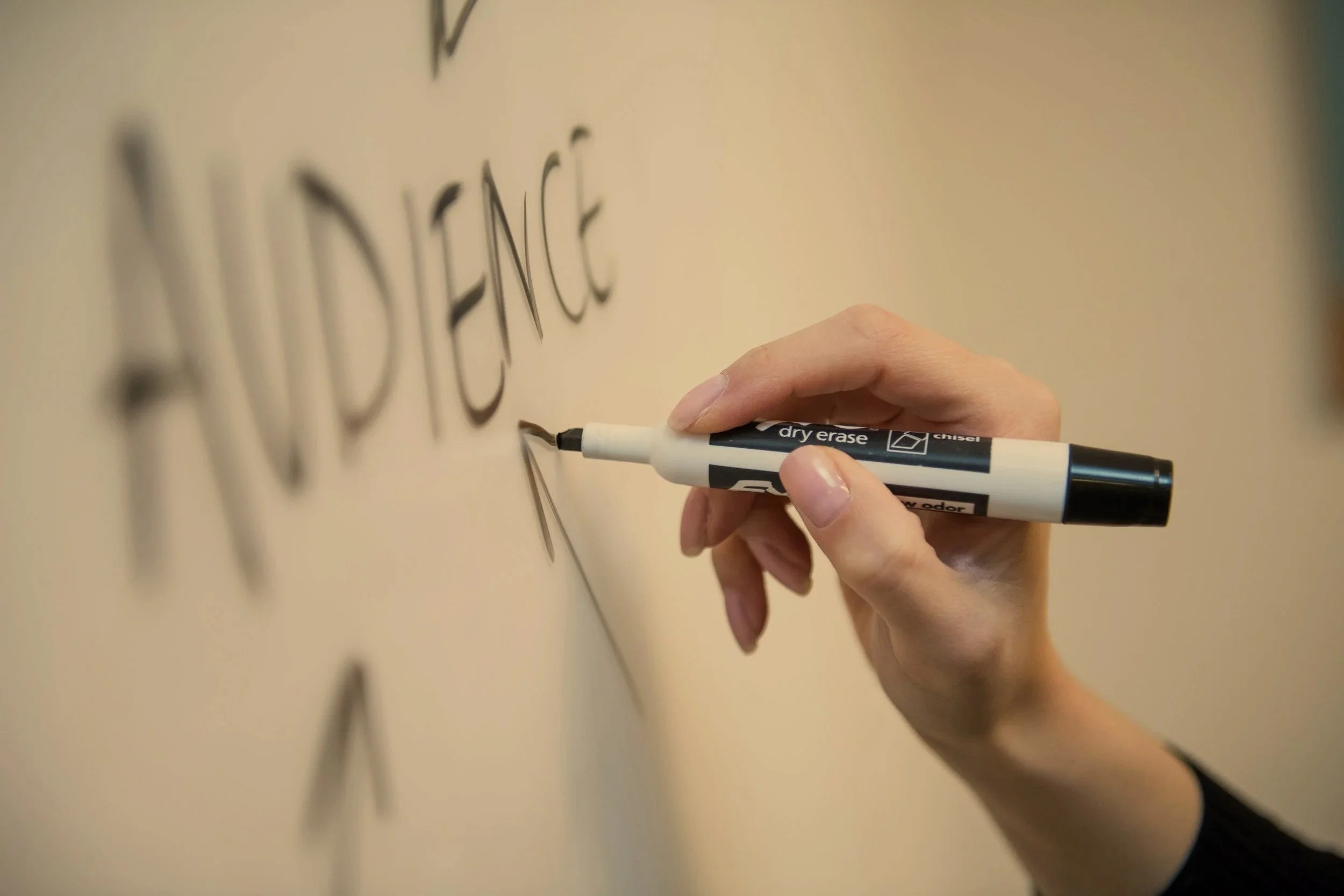 Person writing the word 'AUDIENCE' on a whiteboard with a black marker.