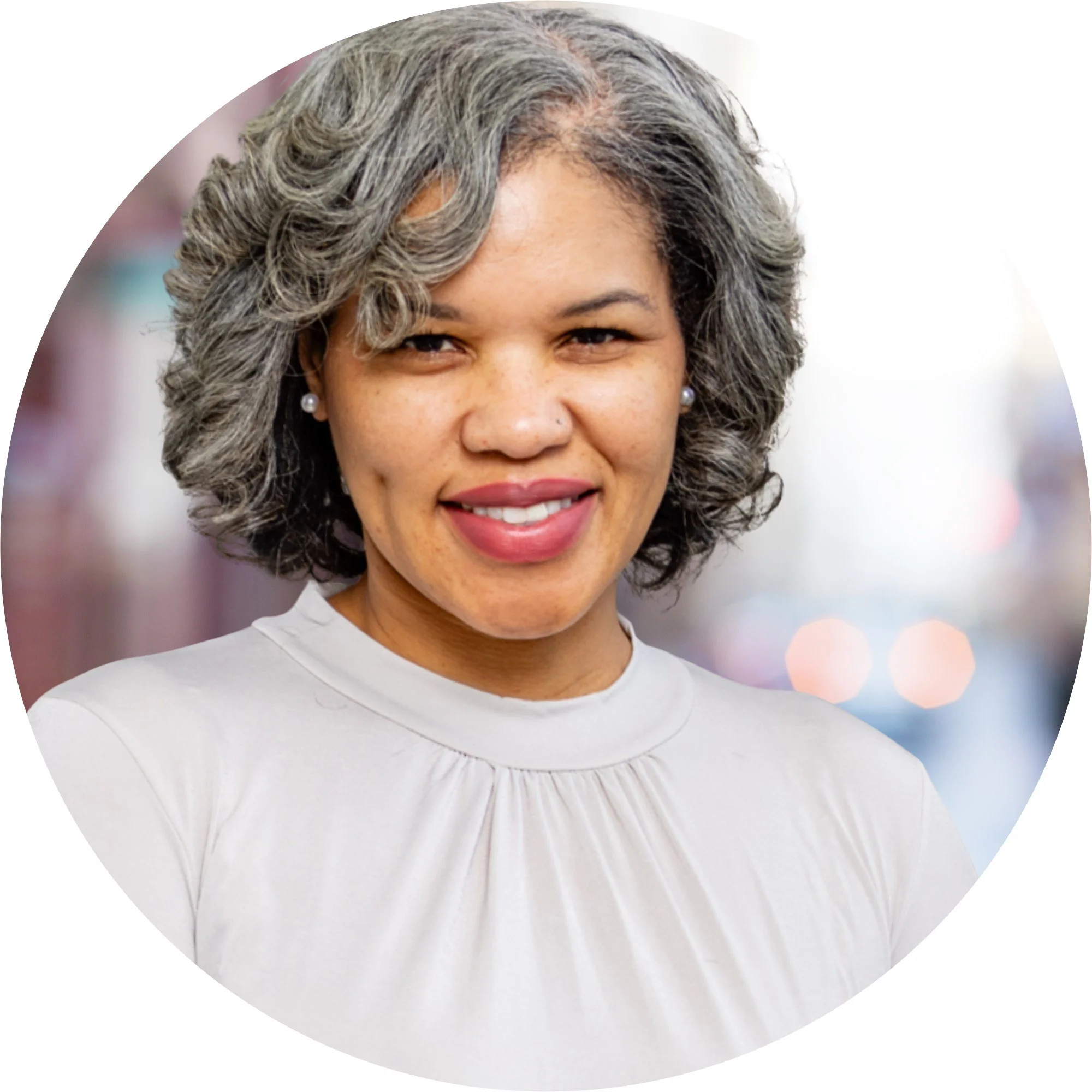 Close-up of a smiling woman with gray curly hair, wearing pearl earrings and a white top, with a blurred city street background.