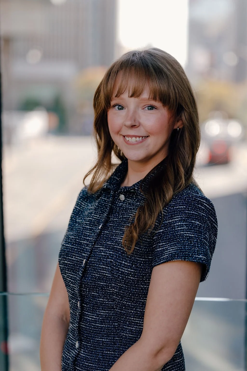 Katelyn Watkins - A young woman with long brown hair and bangs, smiling, standing outdoors with a cityscape background.