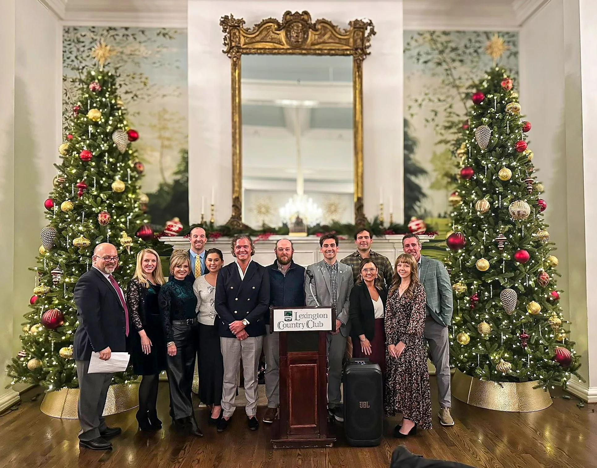 Mortgage Bankers Association of the Bluegrass Board of Directors at a podium in front of Christmas trees and a large mirror in a decorated room, celebrating at the Lexington Country Club.