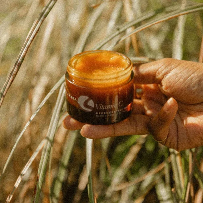 A hand holding an open jar of vitamin C cream outdoors among tall grass.