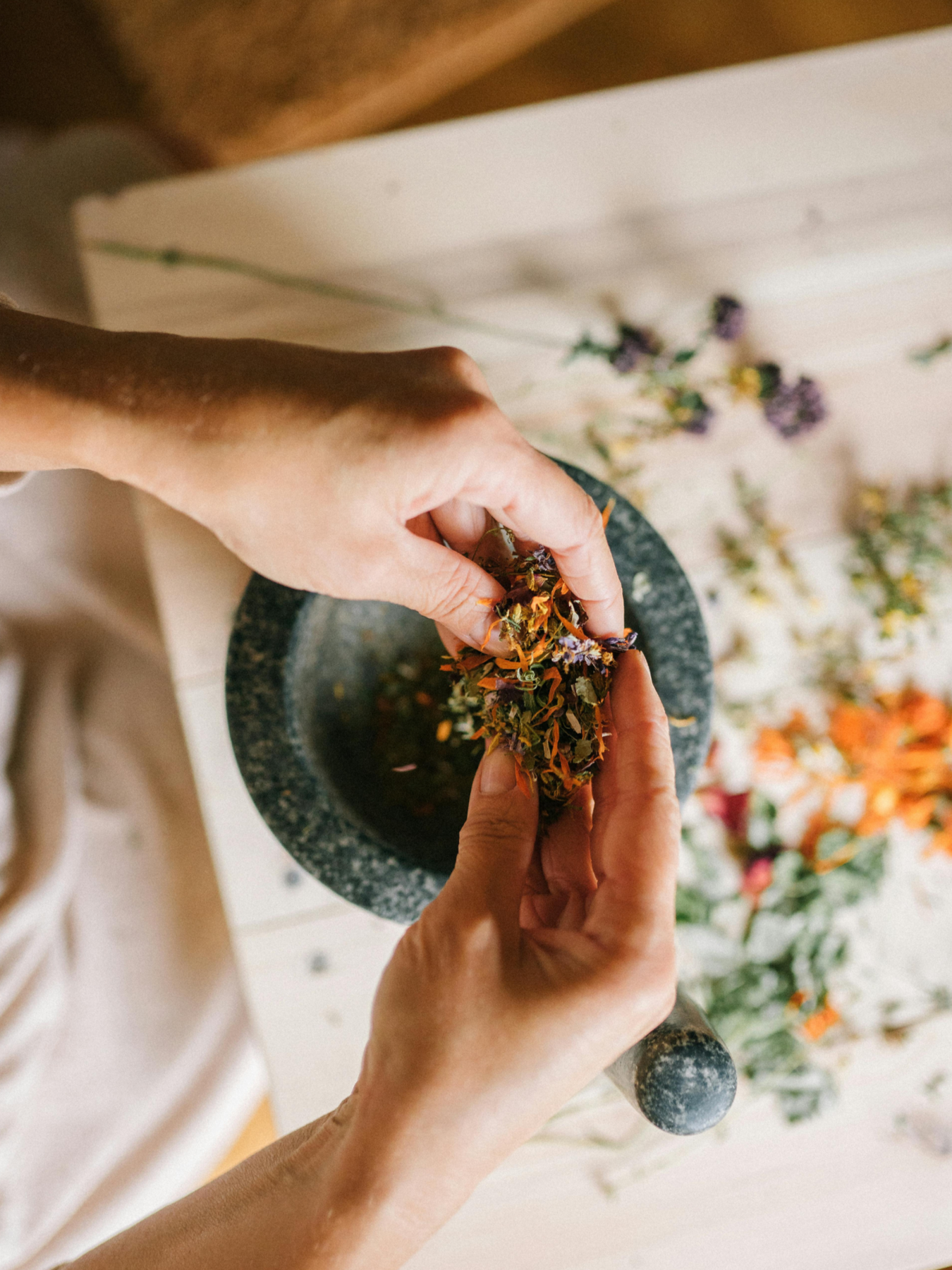 Hands crushing dried herbs or flowers in a mortar and pestle, with dried flowers and herbs scattered on a white surface.