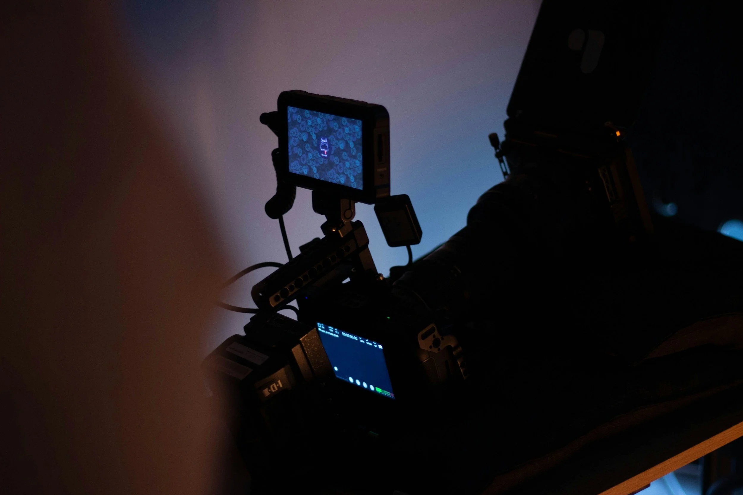 A person operating a professional camera on a tripod, with a small monitor attached, in a dark environment with a gradient blue background.