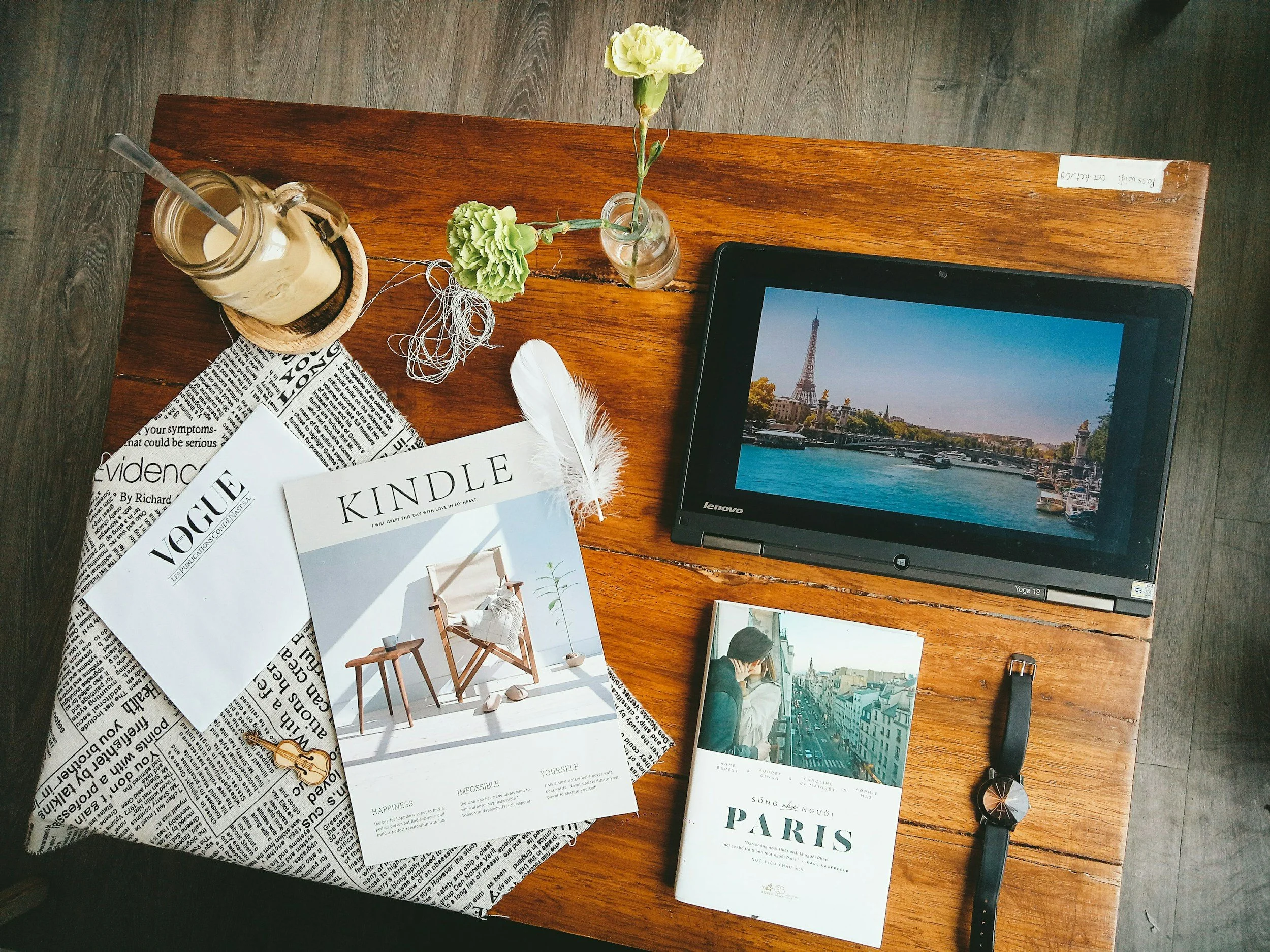 A wooden table holding a laptop displaying a cityscape with the Eiffel Tower, a postcard showing a woman with a city view, a folded newspaper, magazines titled Vogue and Kindle, a wristwatch, a small bag of coffee or tea, a glass jar with a straw, a small vase with yellow and green flowers, and a feather and tangled string on the table.