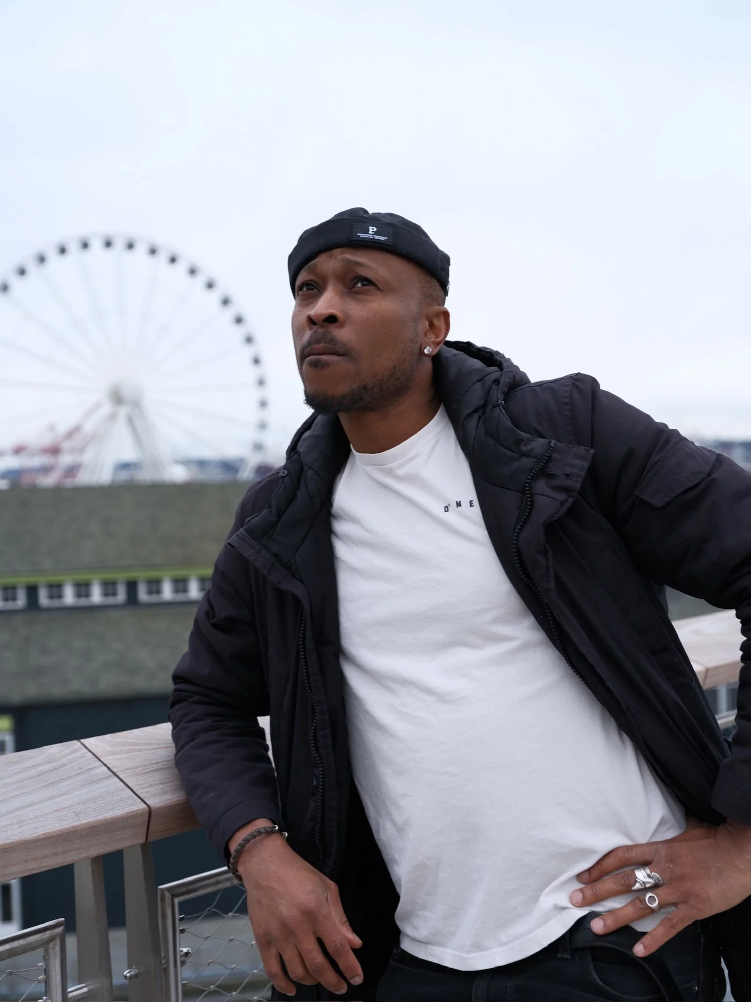 A man wearing a black cap, white t-shirt, and black jacket leaning on a railing with a Ferris wheel in the background, overcast sky.