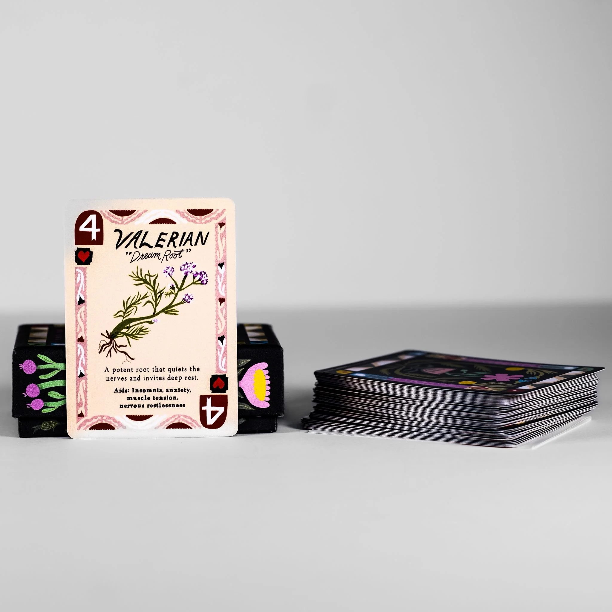 A stack of tarot cards on a white surface with one card upright in front, featuring a botanical illustration and text about Valerian root, with the rest of the deck face down beside it.
