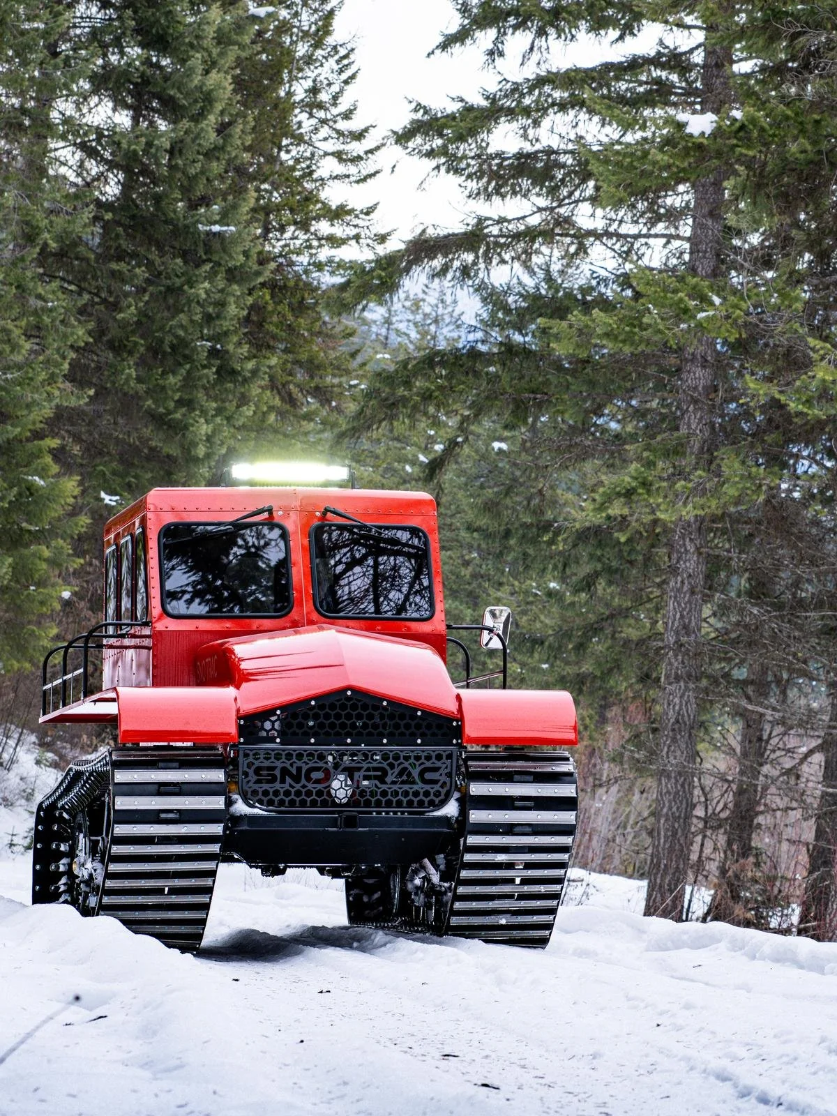 A red snowcat vehicle with large tracks moving through a snow-covered forested area.