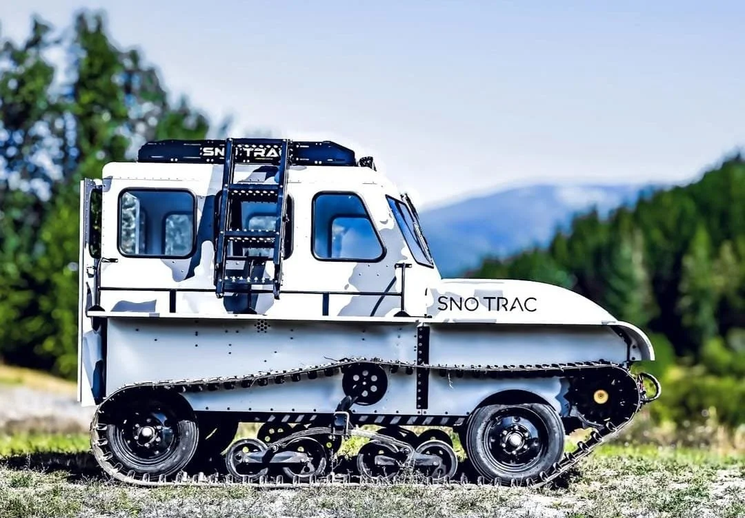 Metal tracked vehicle with a cabin labeled 'SNO TRAC' parked on grass with trees and mountains in the background.