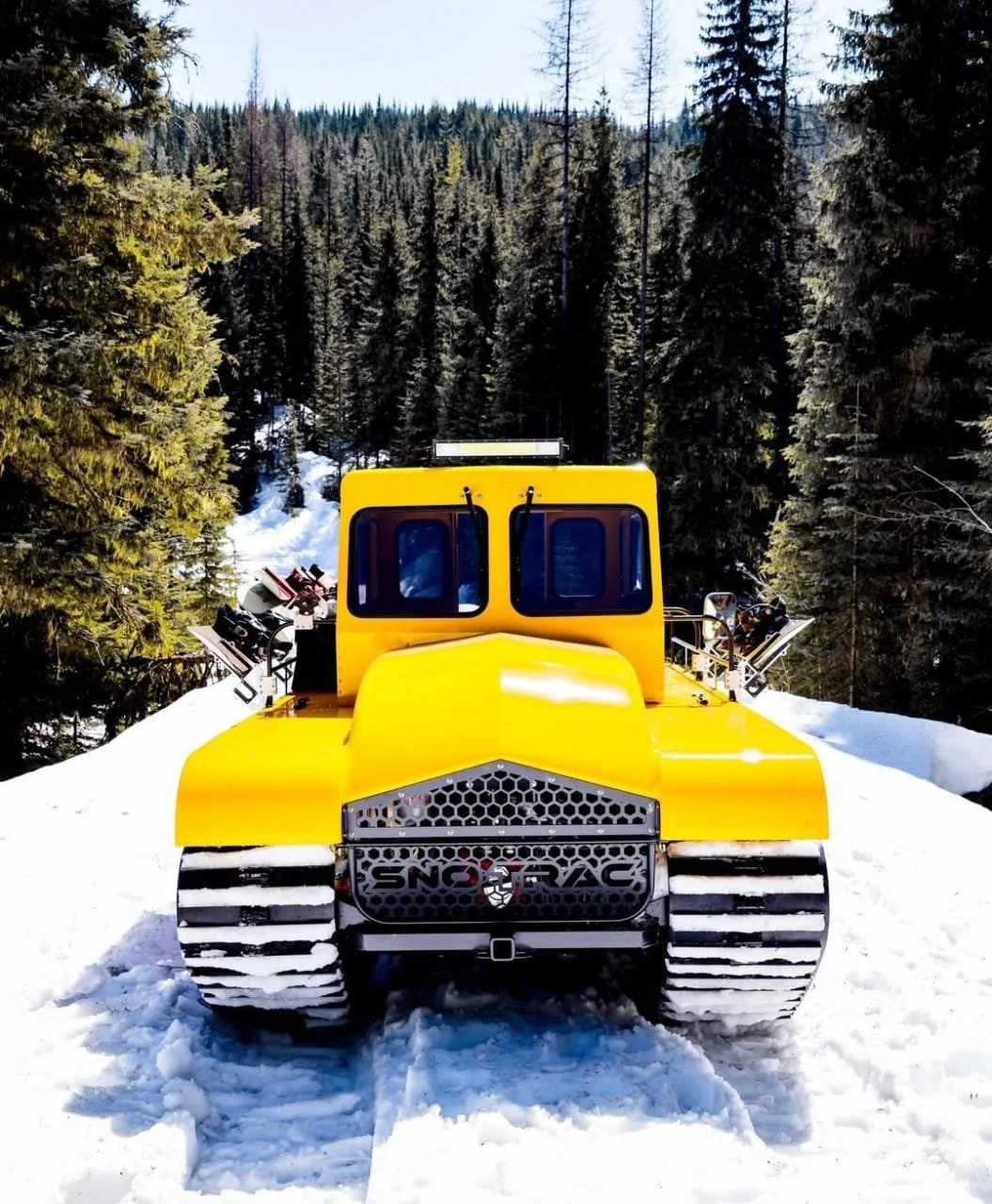 Yellow snow groomer vehicle on snowy trail in a forested area.