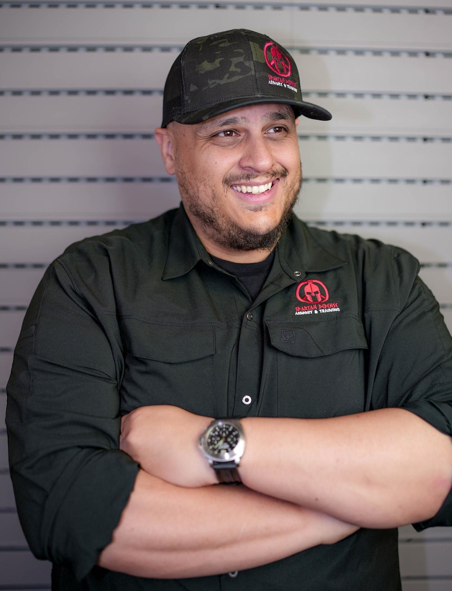 Teddy Collins, Candidate for Colorado State Senate District 4 smiling with arms crossed, wearing a black shirt with red logo and a camouflage baseball cap, standing in front of a beige striped wall.