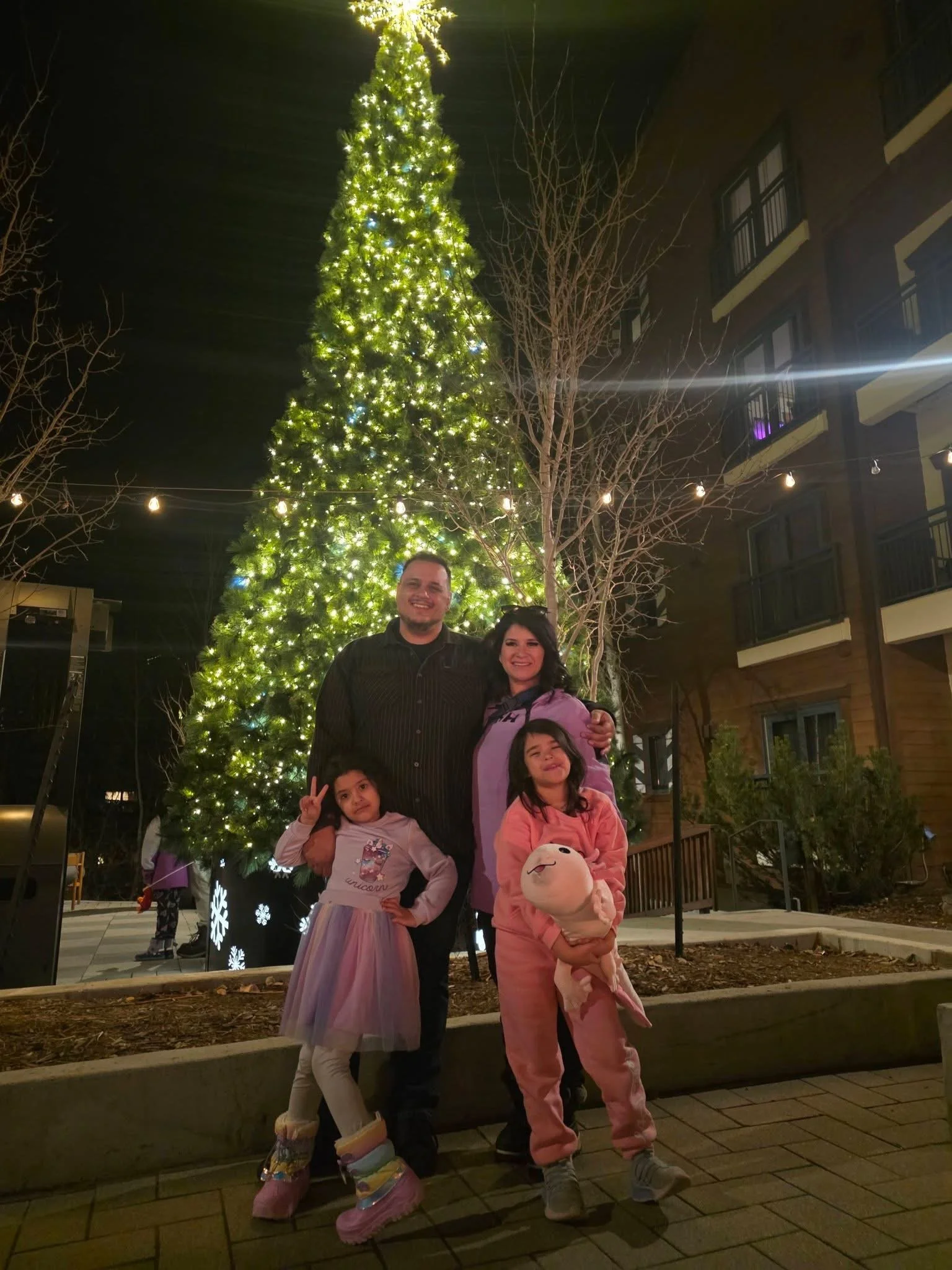 Teddy Collins, Candidate for Colorado SD 4 with his family posing in front of a decorated Christmas tree with lights at night outside a building.