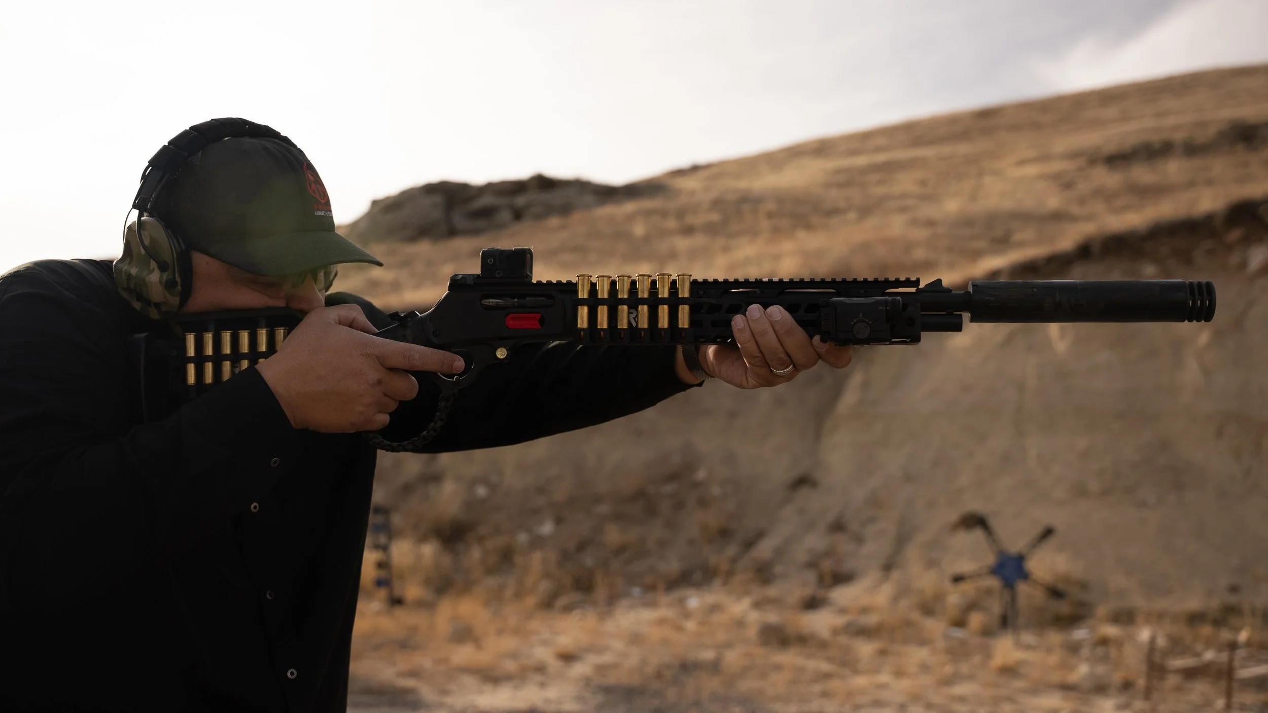 Teddy Collins, Candidate for Colorado State Senate District 4 aiming a rifle with a suppressor and a scope at a shooting range outdoors in a mountainous area.