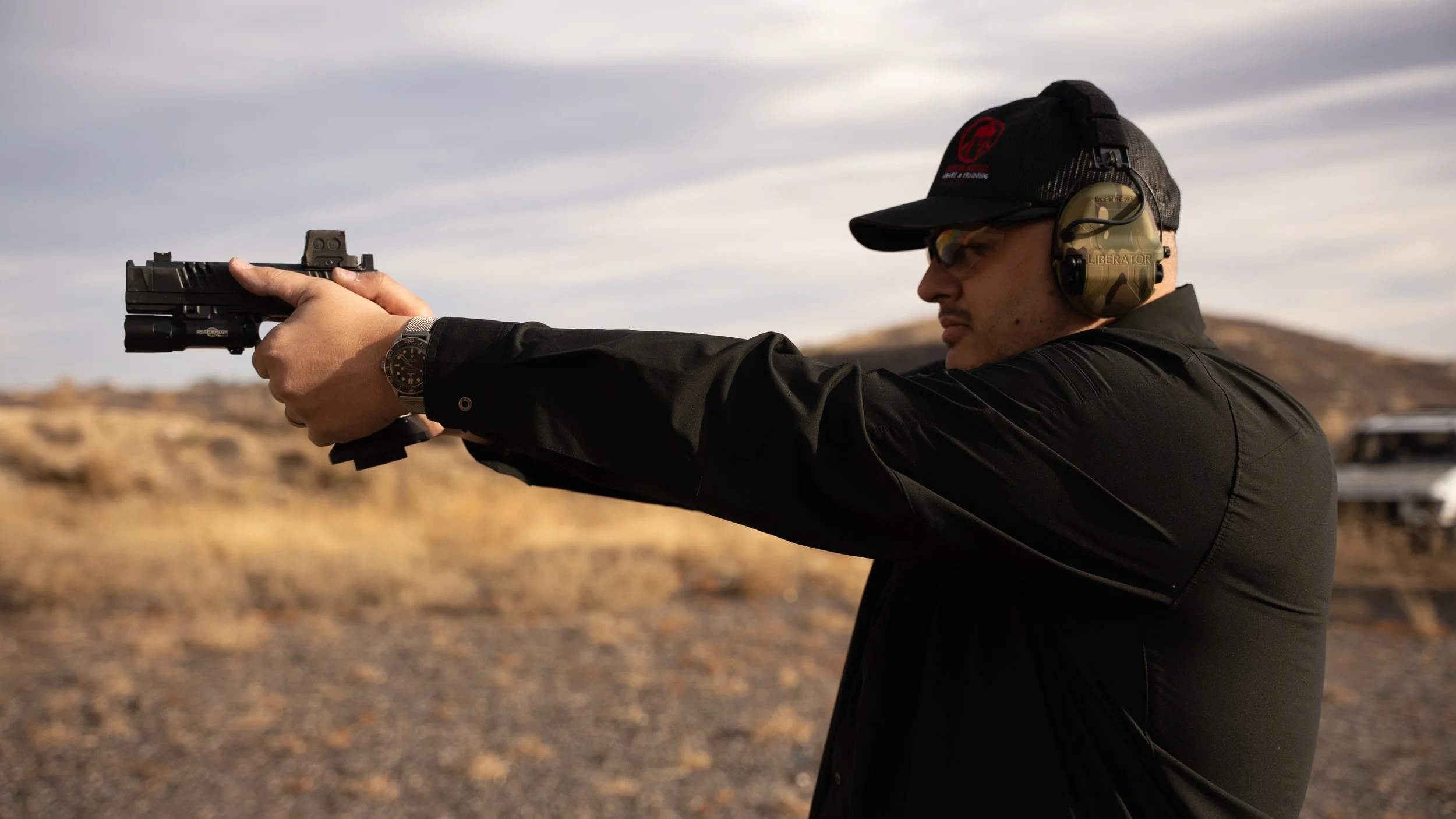 Teddy Collins, Candidate for Colorado State Senate District 4 shooting a handgun outdoors in a desert landscape, wearing a hat, glasses, and headphones for ear protection.