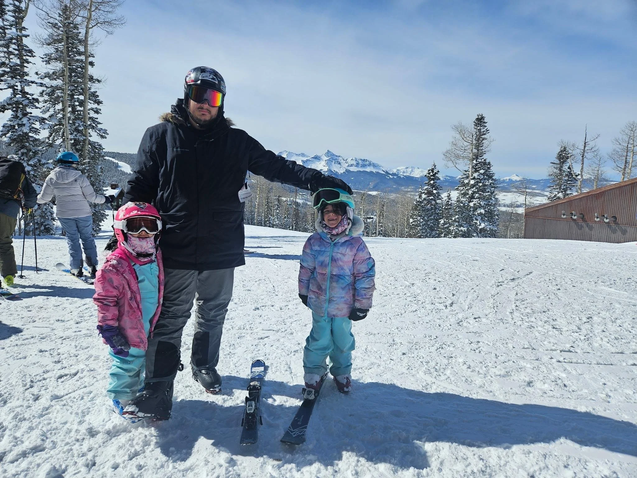 Teddy Collins, Candidate for Colorado State Senate District 4 and his daughters in ski gear on a snowy mountain, with ski equipment and other skiers in the background, mountain peaks in the distance under a clear blue sky.