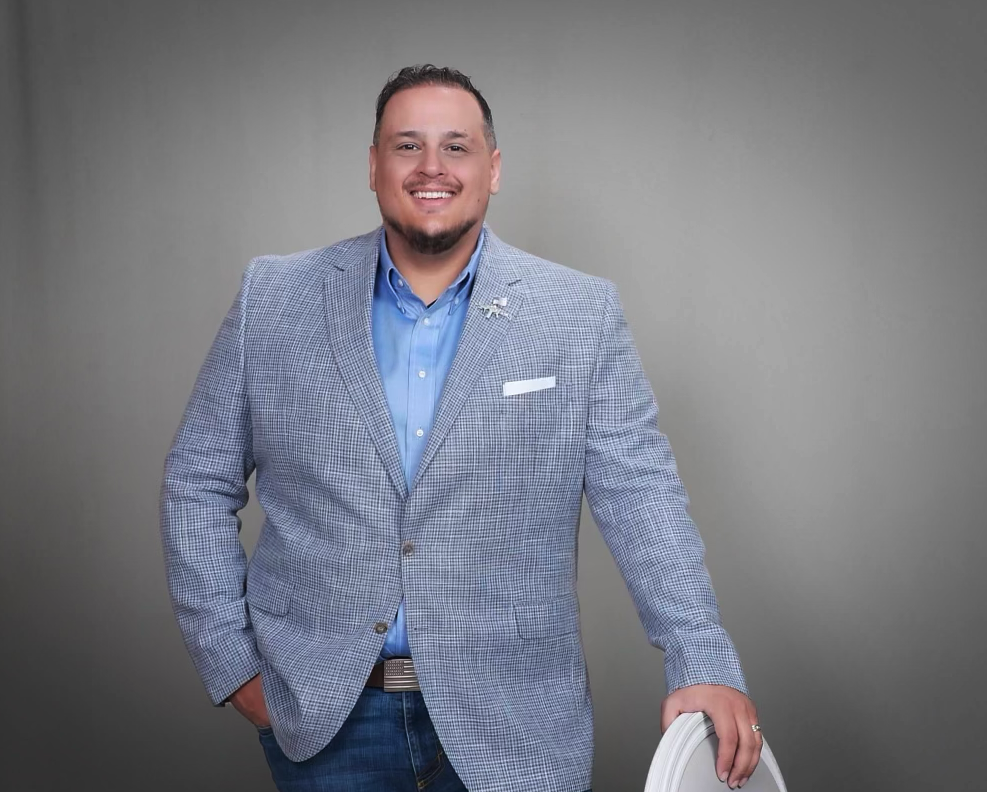 Teddy Collins, Candidate for Colorado SD 4 in a light gray checked blazer and blue shirt, standing against a plain gray background.