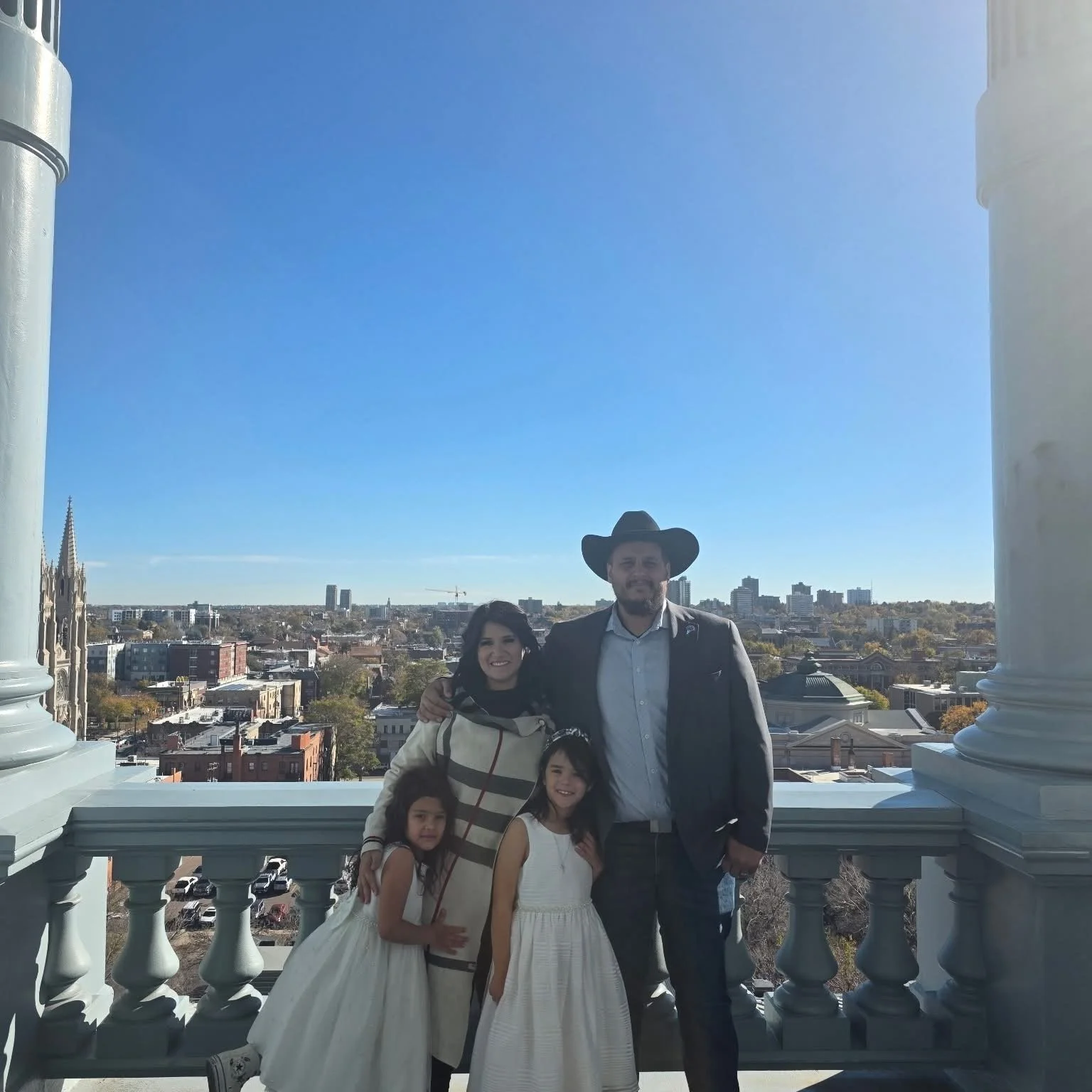 Teddy Collins, Candidate for Colorado SD 4 with his family standing on a balcony in the Colorado Capitol with Denver in the background during daytime. 