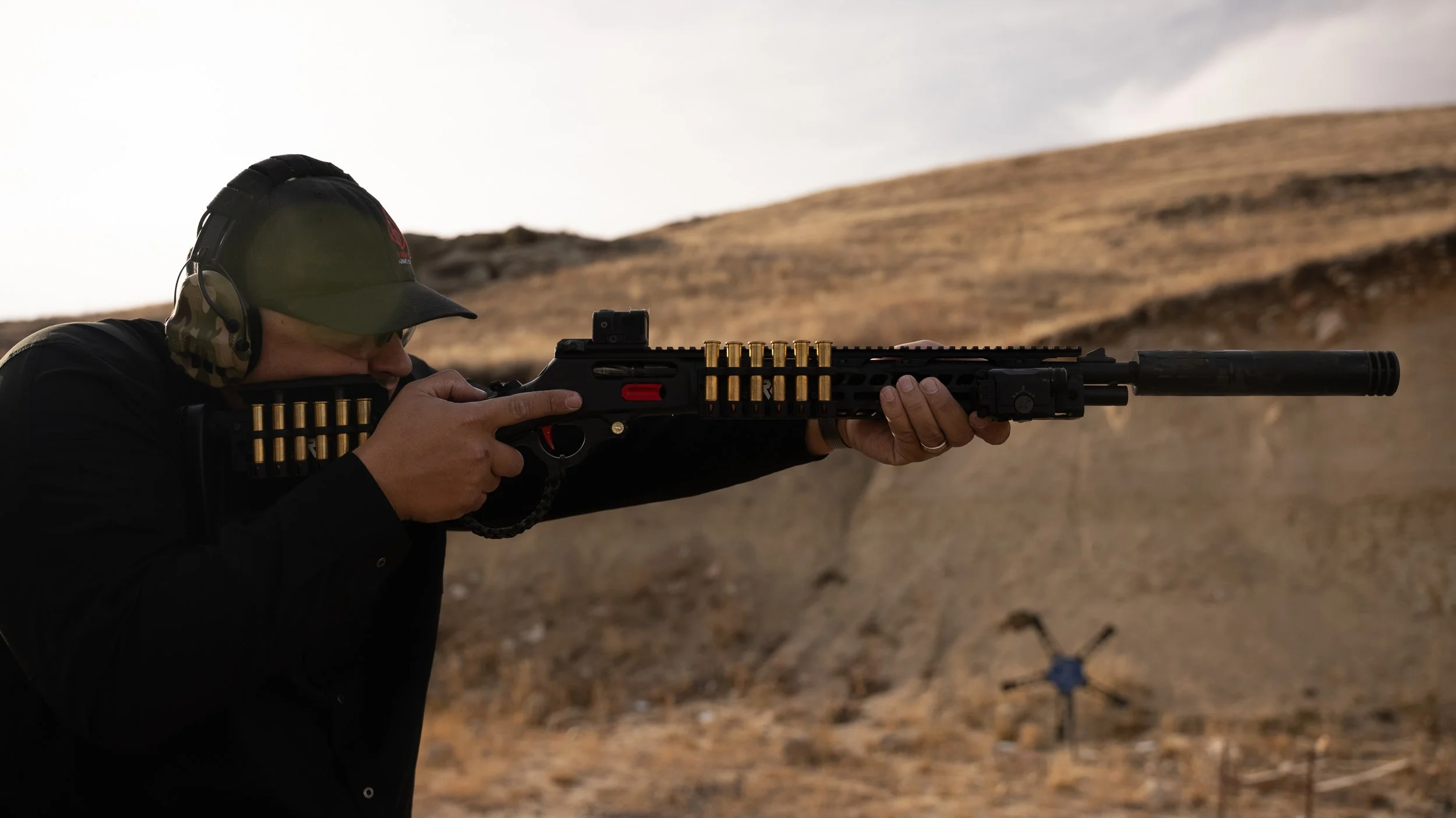 Teddy Collins, Candidate for Colorado State Senate District 4 aiming a rifle with a silencer in a desert landscape, wearing headphones and a cap.
