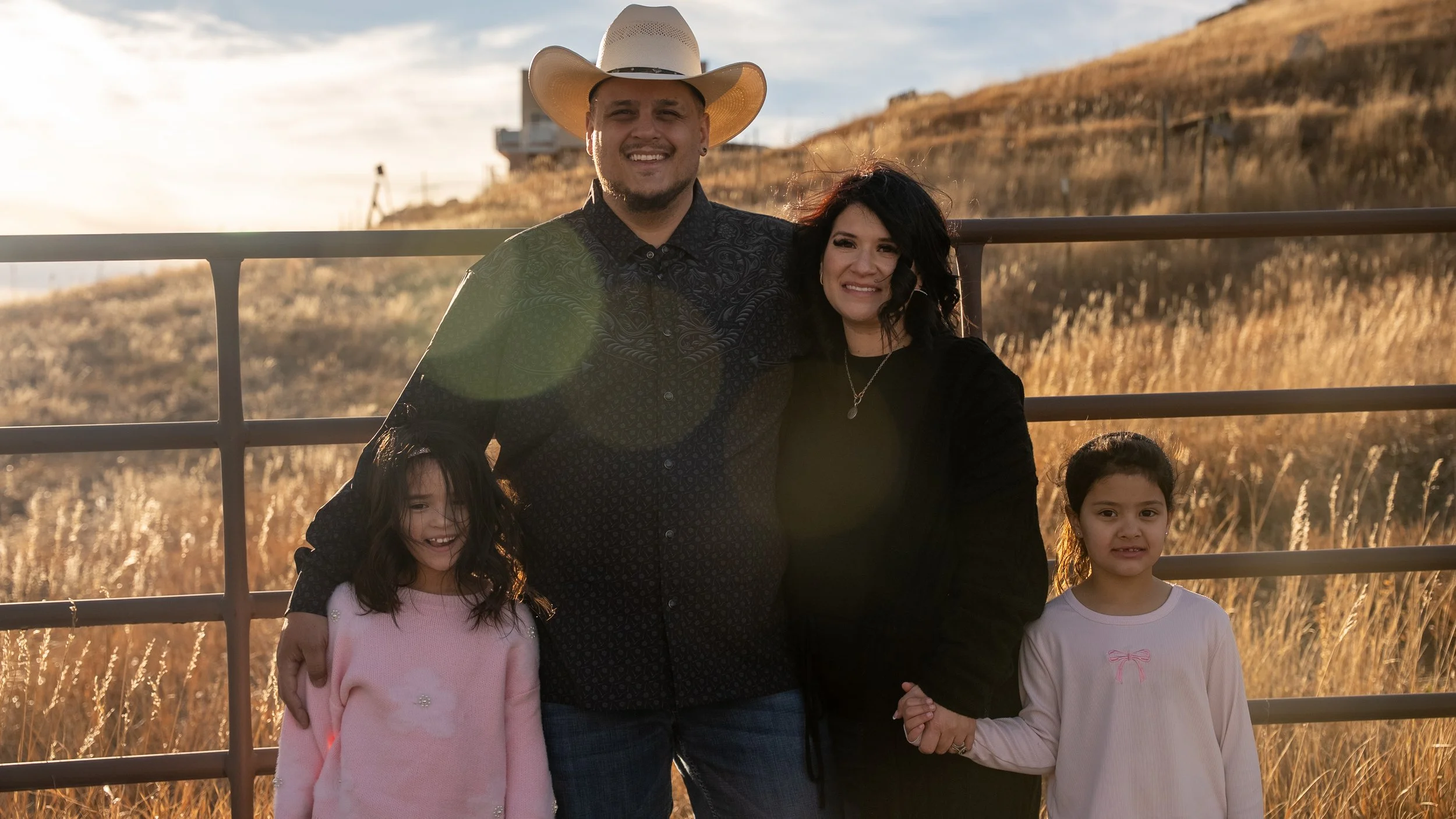 Teddy Collins, Candidate for Colorado State Senate District 4 and his family of four standing outdoors in front of a metal fence with tall grass and hills in the background during golden hour. 