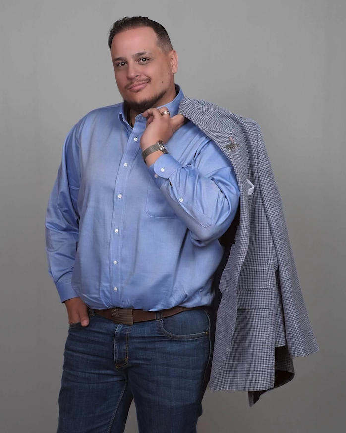Teddy Collins, Candidate for Colorado SD 4 in a blue button-up shirt and jeans holding a blazer over his shoulder, standing against a plain gray background.