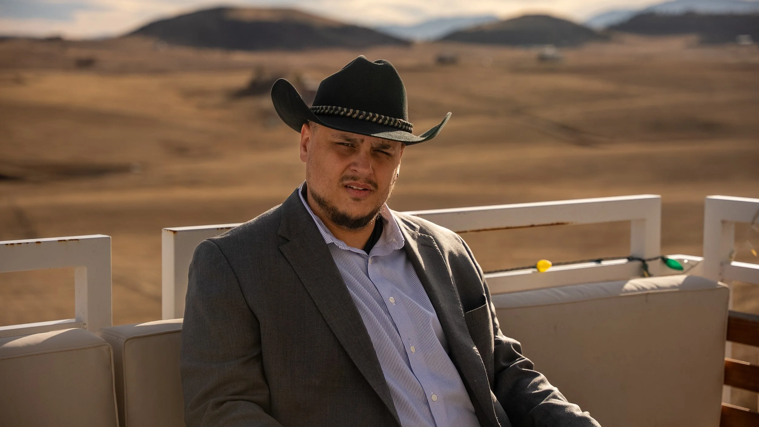 Teddy Collins, Candidate for Colorado State Senate District 4 wearing a black cowboy hat, gray blazer, and striped shirt, sitting outdoors with a muddy, hilly landscape in the background.