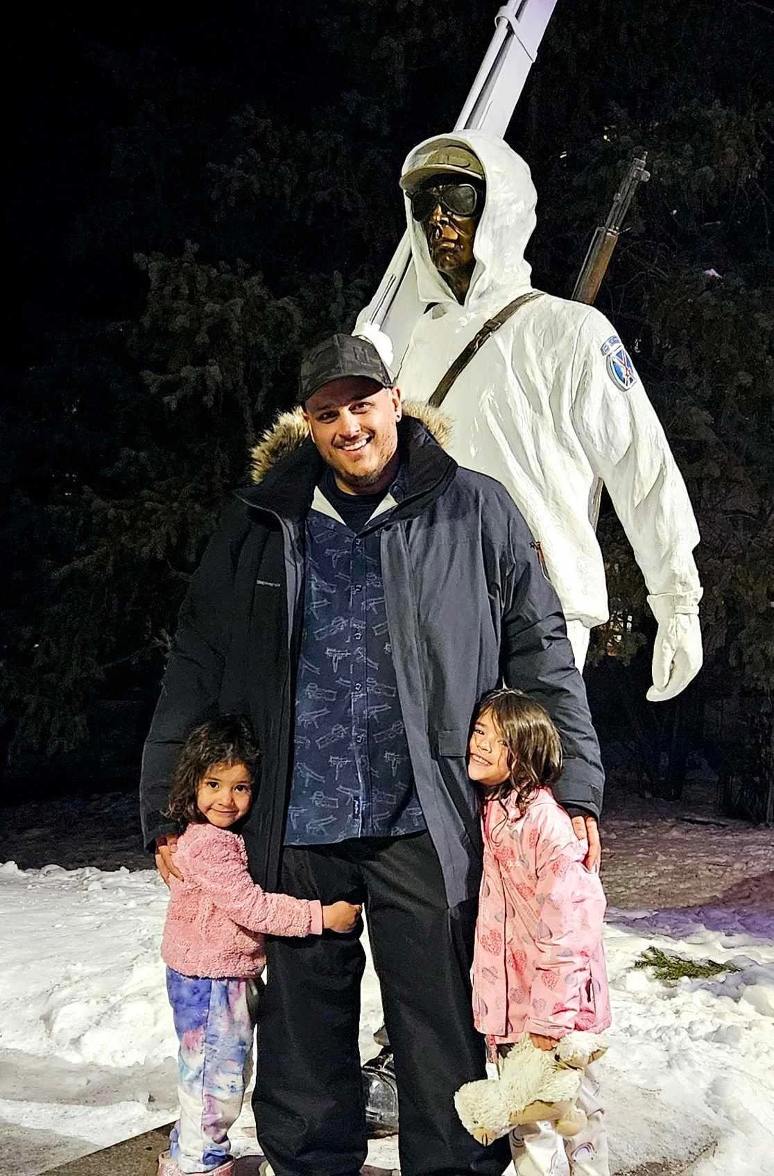 Teddy Collins, Candidate for Colorado State Senate District 4 and his two daughters smiling and hugging in front of a large white statue of a soldier in a snowy outdoor setting at night.