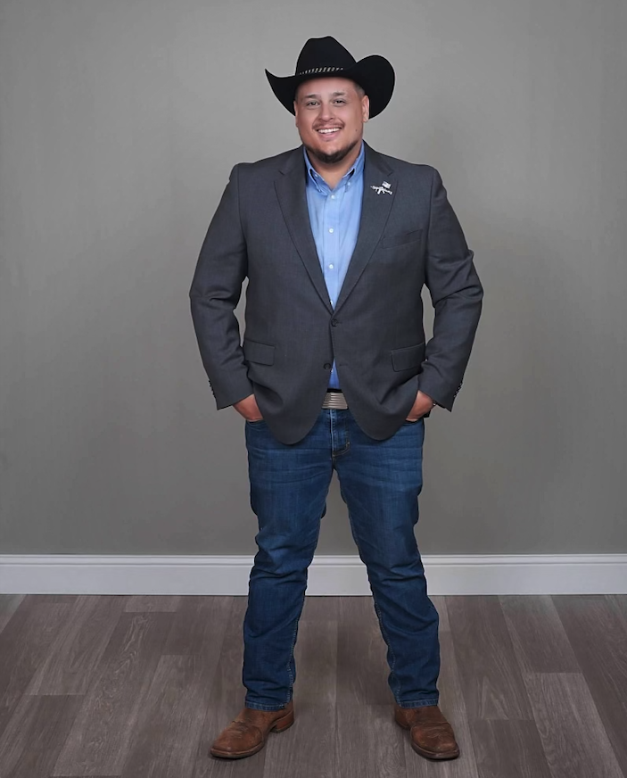 Teddy Collins, Candidate for Colorado SD 4 wearing a gray suit jacket, blue shirt, jeans, brown boots, and a black cowboy hat, standing with hands in pockets against a plain gray background.