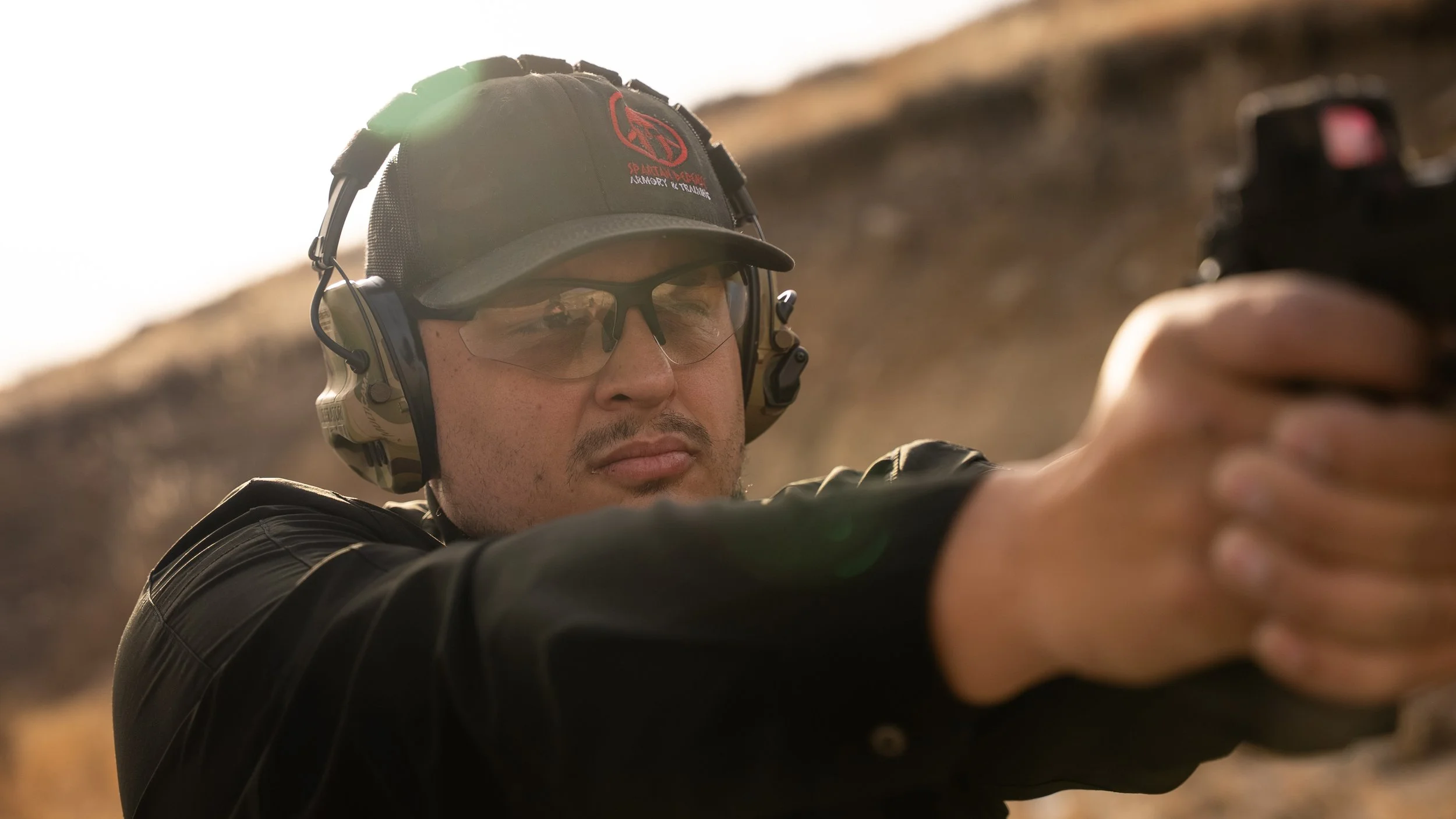 Teddy Collins, Candidate for Colorado State Senate District 4 at outdoor shooting range aiming a handgun with safety glasses, earmuffs, and a cap.