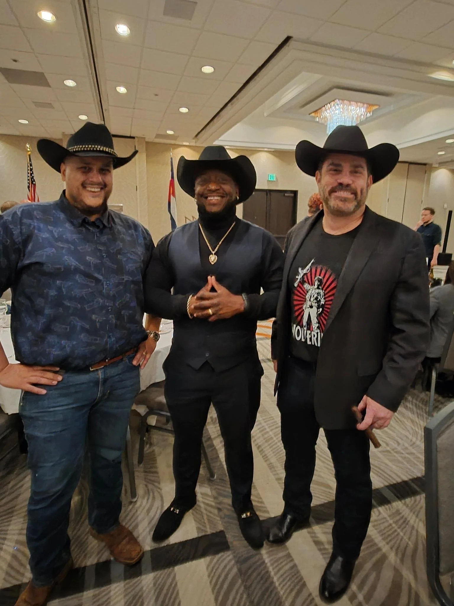 Teddy Collins, Candidate for Colorado State Senate District 4 and two other men wearing cowboy hats standing together at an indoor event, with flags and people in the background.