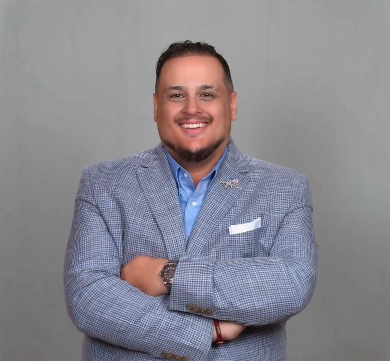 Teddy Collins, Candidate for Colorado SD 4, smiling with arms crossed, dressed in a light gray checked blazer, blue dress shirt, standing against a plain gray background.