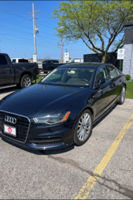 A black Audi sedan parked in a parking lot next to a yellow handicap parking space line, with trees and utility poles in the background.