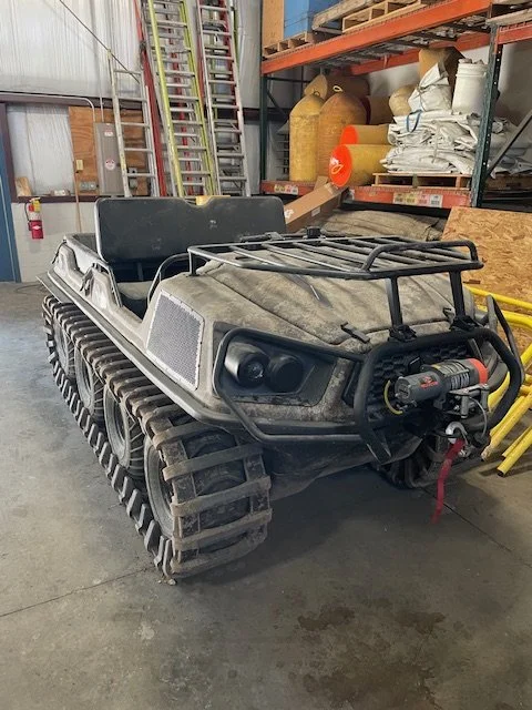 A tracked all-terrain vehicle with a black front guard, located inside a warehouse or storage area, with shelves and supplies in the background.