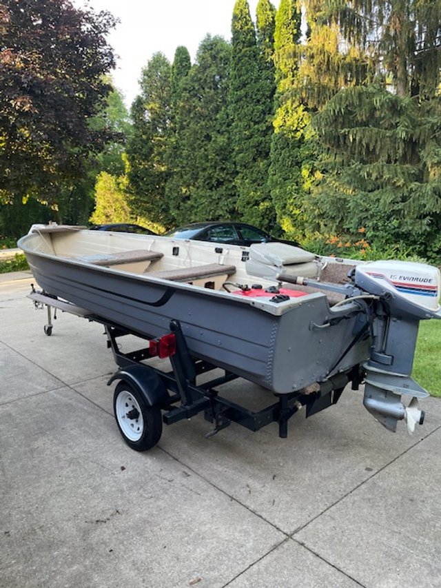 A small boat on a trailer parked on a driveway with green trees and cars in the background.