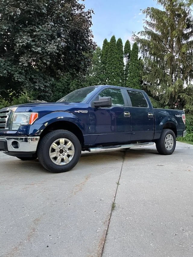 Blue Ford F-150 pickup truck parked on a concrete driveway with trees in the background.