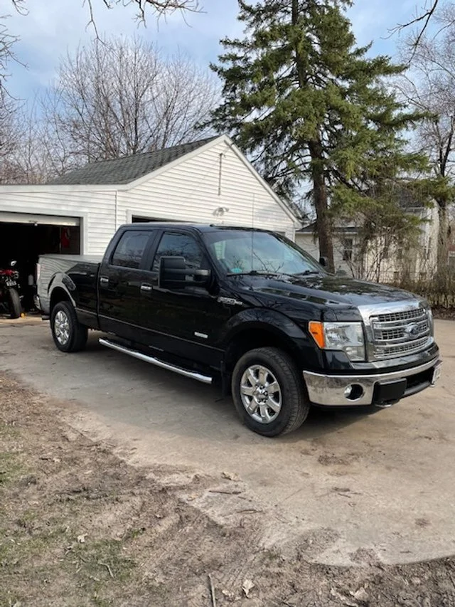Black Ford pickup truck parked on a dirt driveway in front of a white garage, with trees and a house in the background.